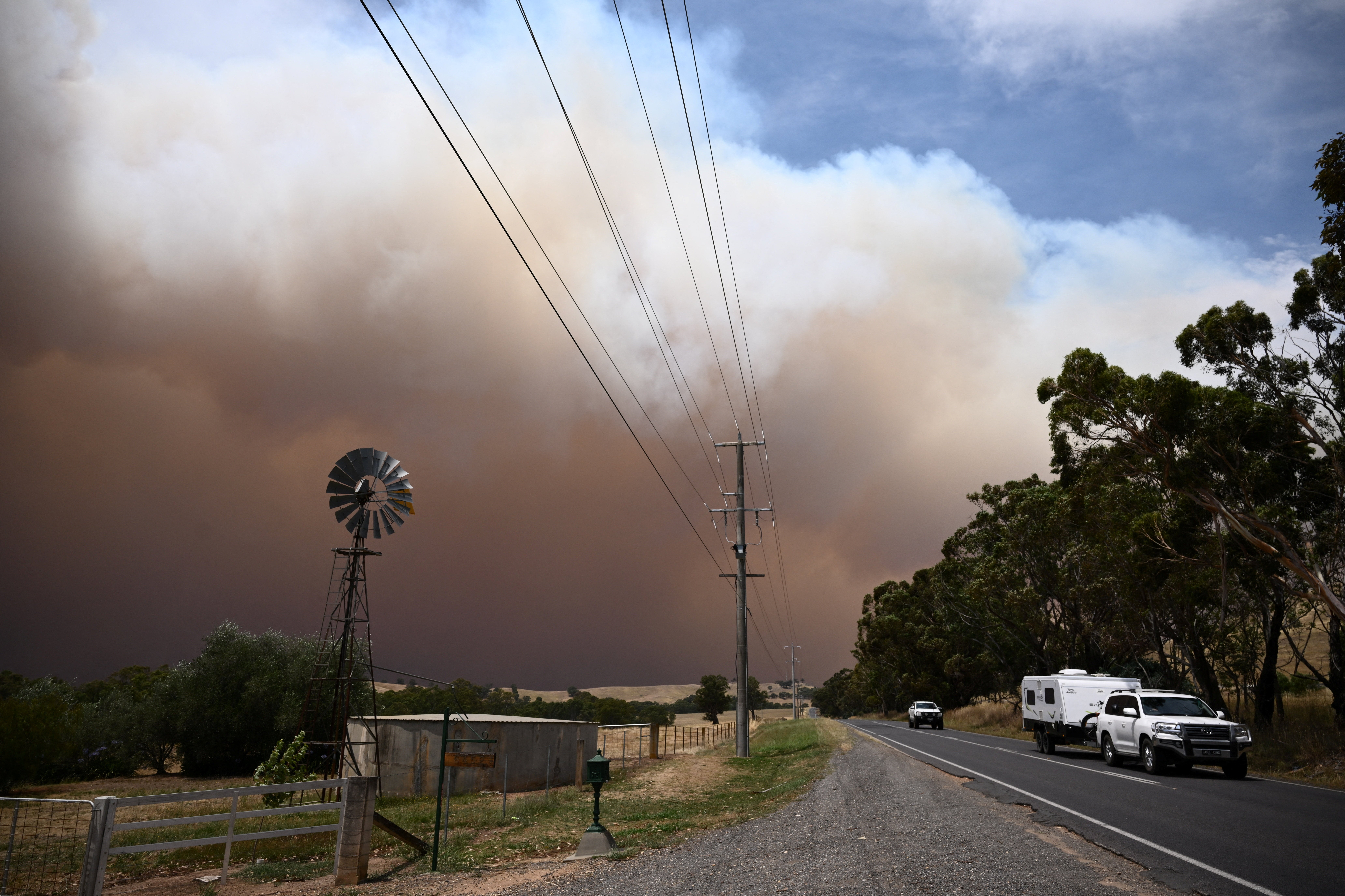 <p>A car towing a caravan drives away from a plume of smoke from the Longwood bushfire, between Seymour and Yea, as out-of-control fires burn across Victoria</p>