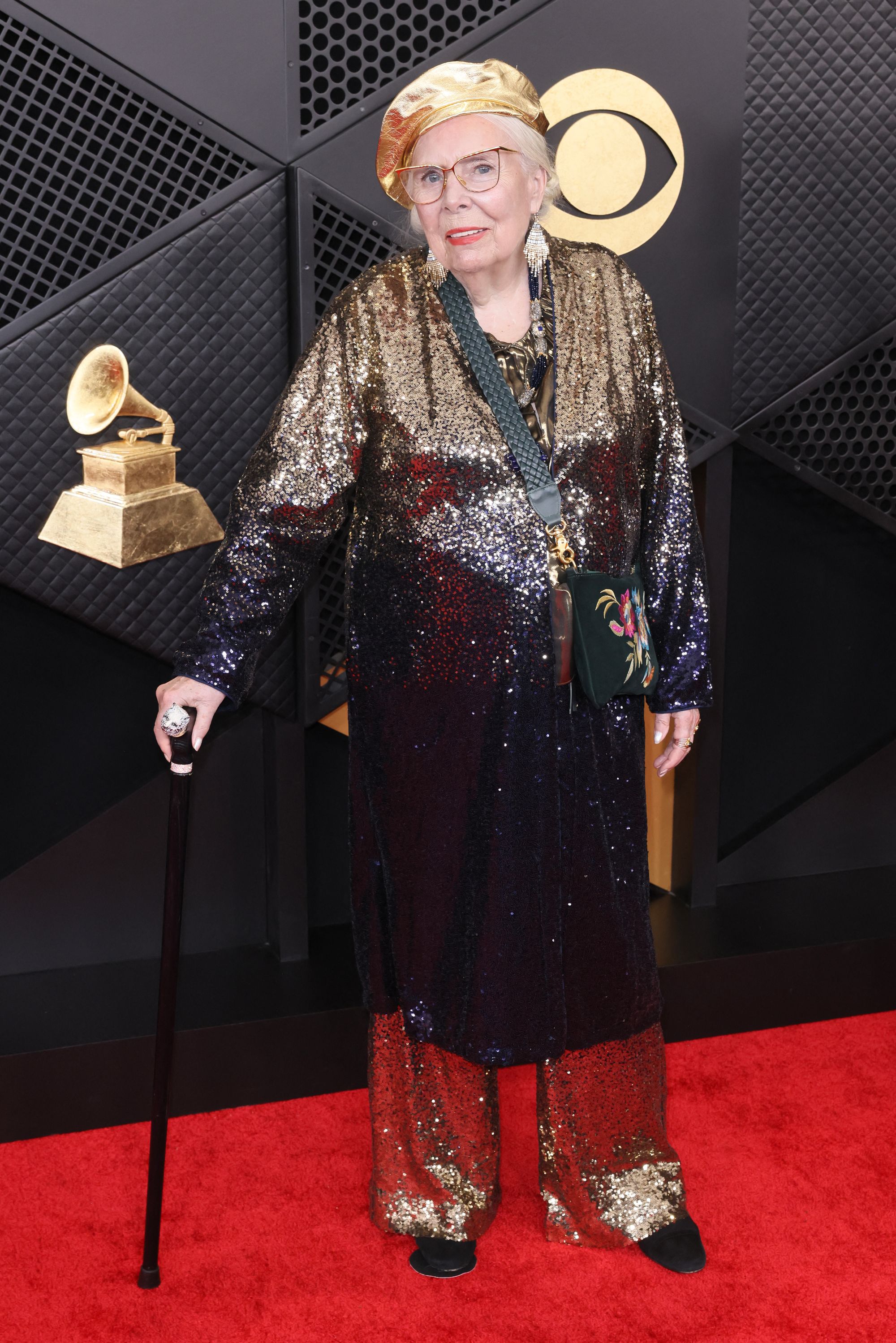 Joni Mitchell took over the red carpet in all sparkles, wearing gold flare pants and a matching long-sleeved caftan-type top. She paired the look with a black floral cross-body purse