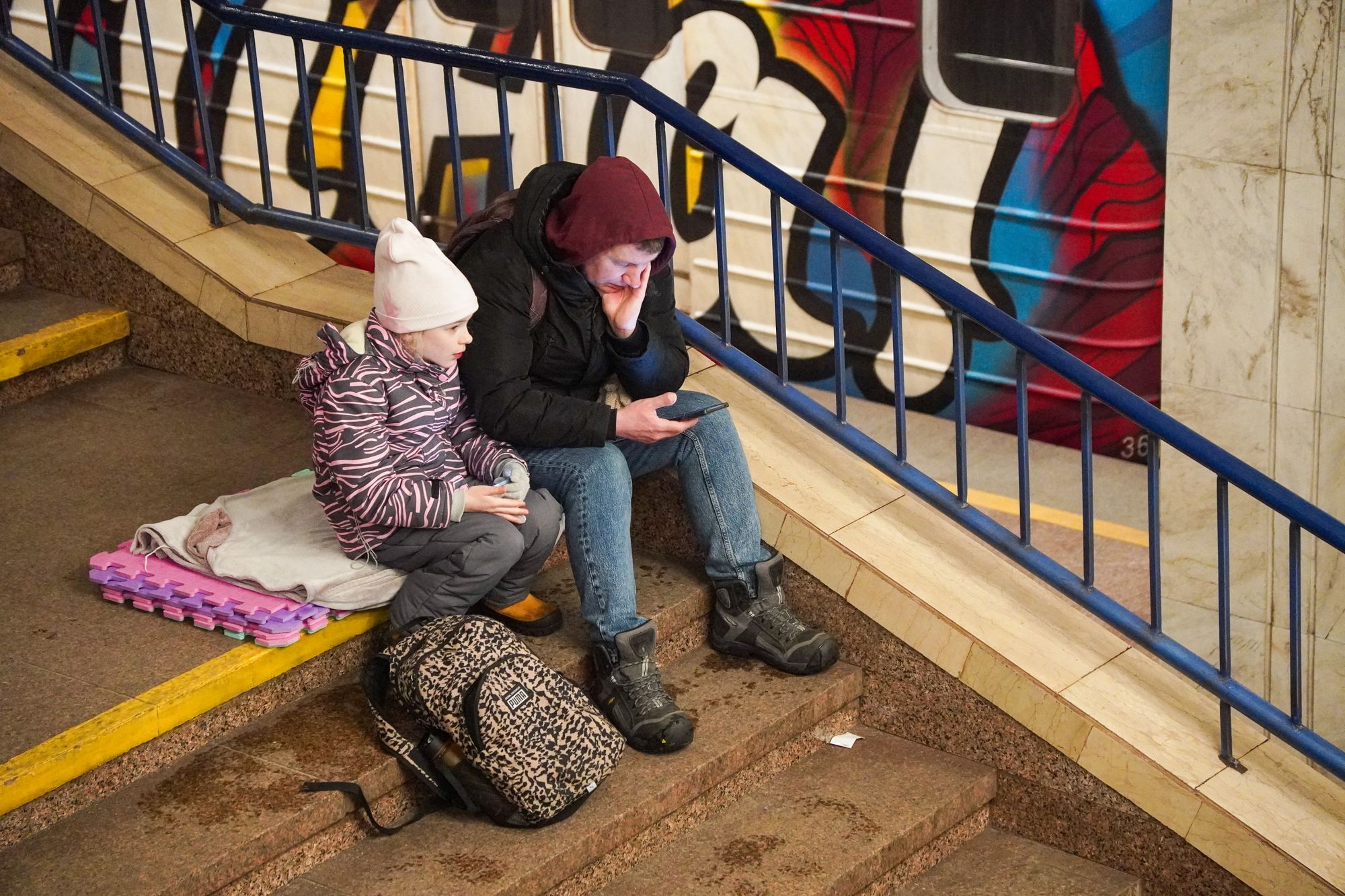 A man checks his phone while taking shelter with a child at a metro station during an air raid alert in Kyiv