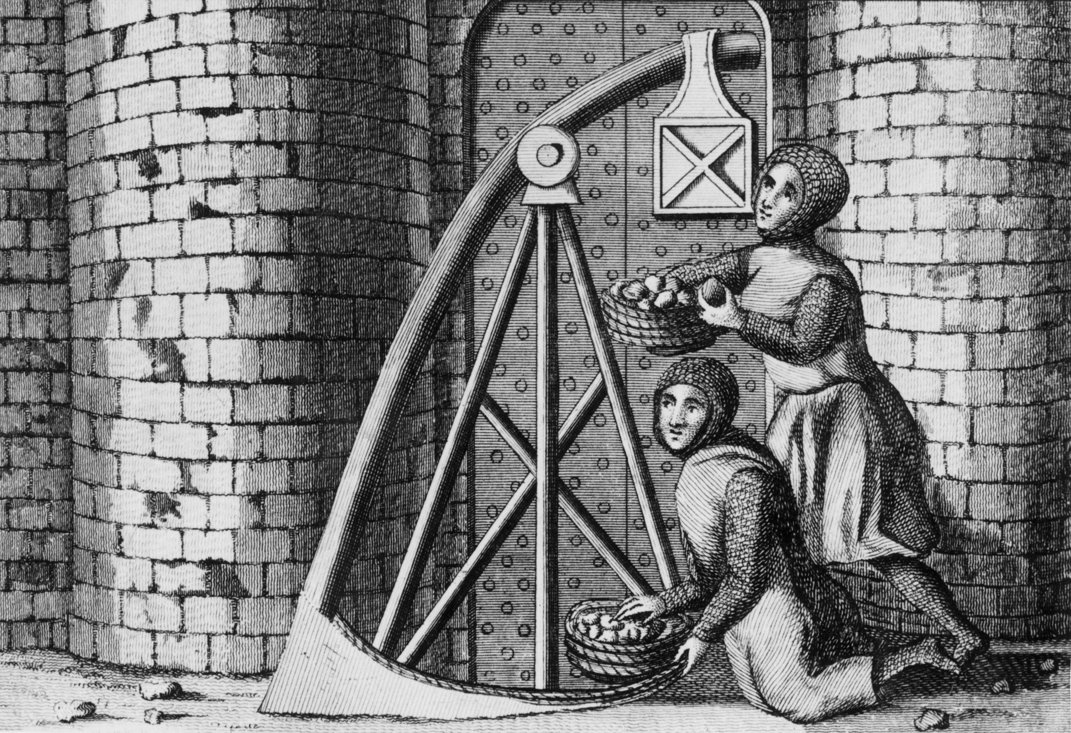 Soldiers wearing chain mail load baskets of stones on to a trebuchet behind the walls of a castle circa 1400