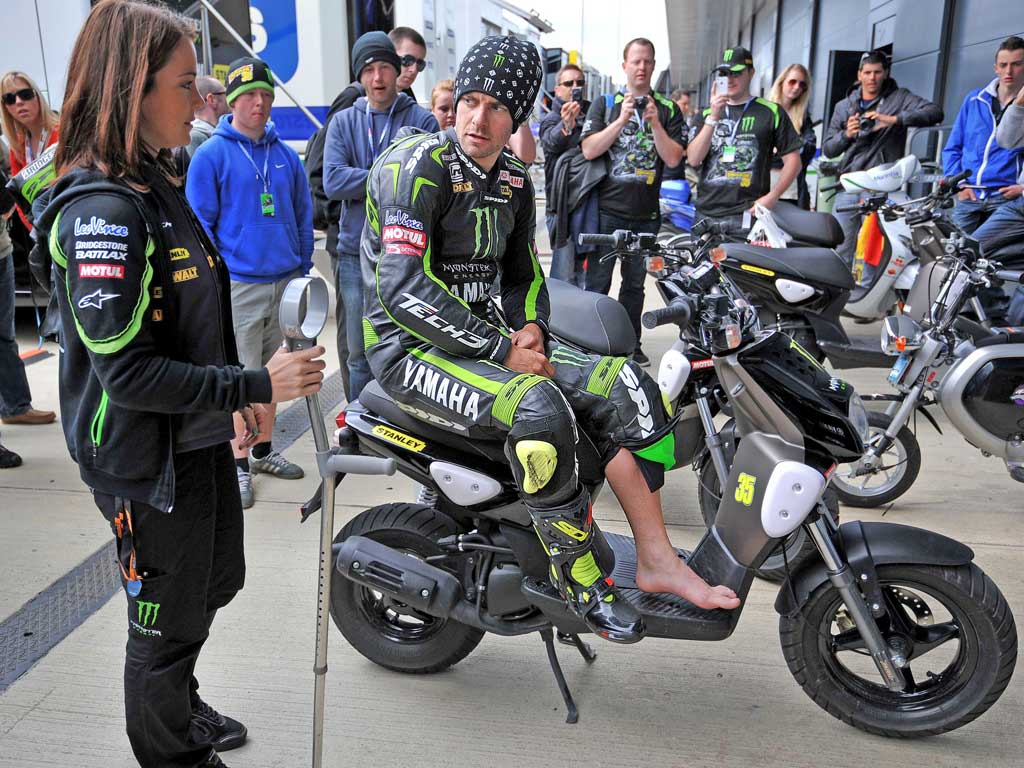 Cal Crutchlow sits on a scooter as a team member holds his crutch ahead of the British Grand Prix at Silverstone