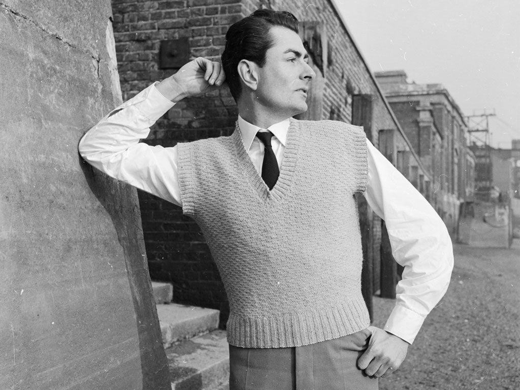 13th January 1958: A man wearing a woollen V-neck pullover accentuating his shirt and tie leans ostentatiously on the wall of a back street. Woman's Illustration