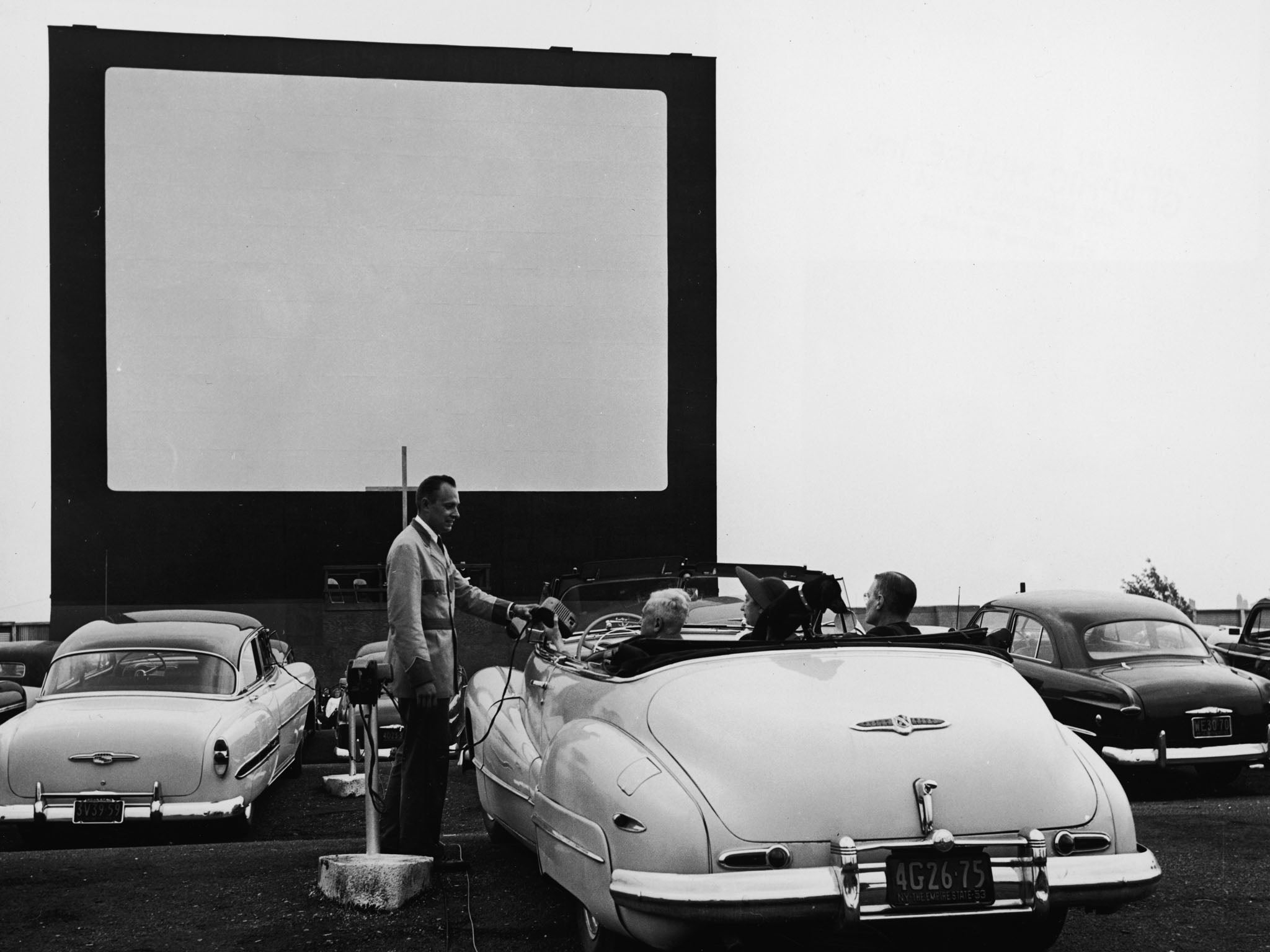 A uniformed drive-in theater attendant hands a clip-on speaker to the driver of convertible while the car's other passengers watch, New York, early 1950s.