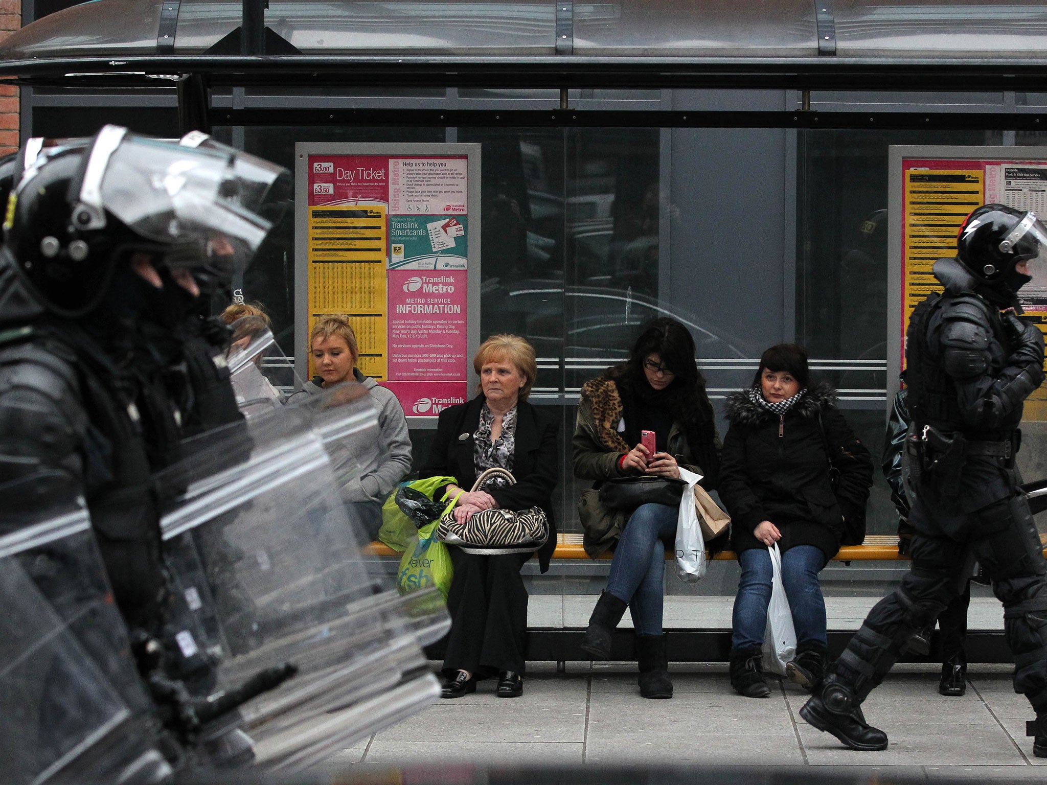 People wait at a bus stop as police dressed in riot gear walk by as loyalists flying march outside Belfast City Hall in protest over Belfast city council's decision to restrict the number of days the British Union Flag can be flown over the city hall in Belfast, Northern Ireland on January 5, 2013.