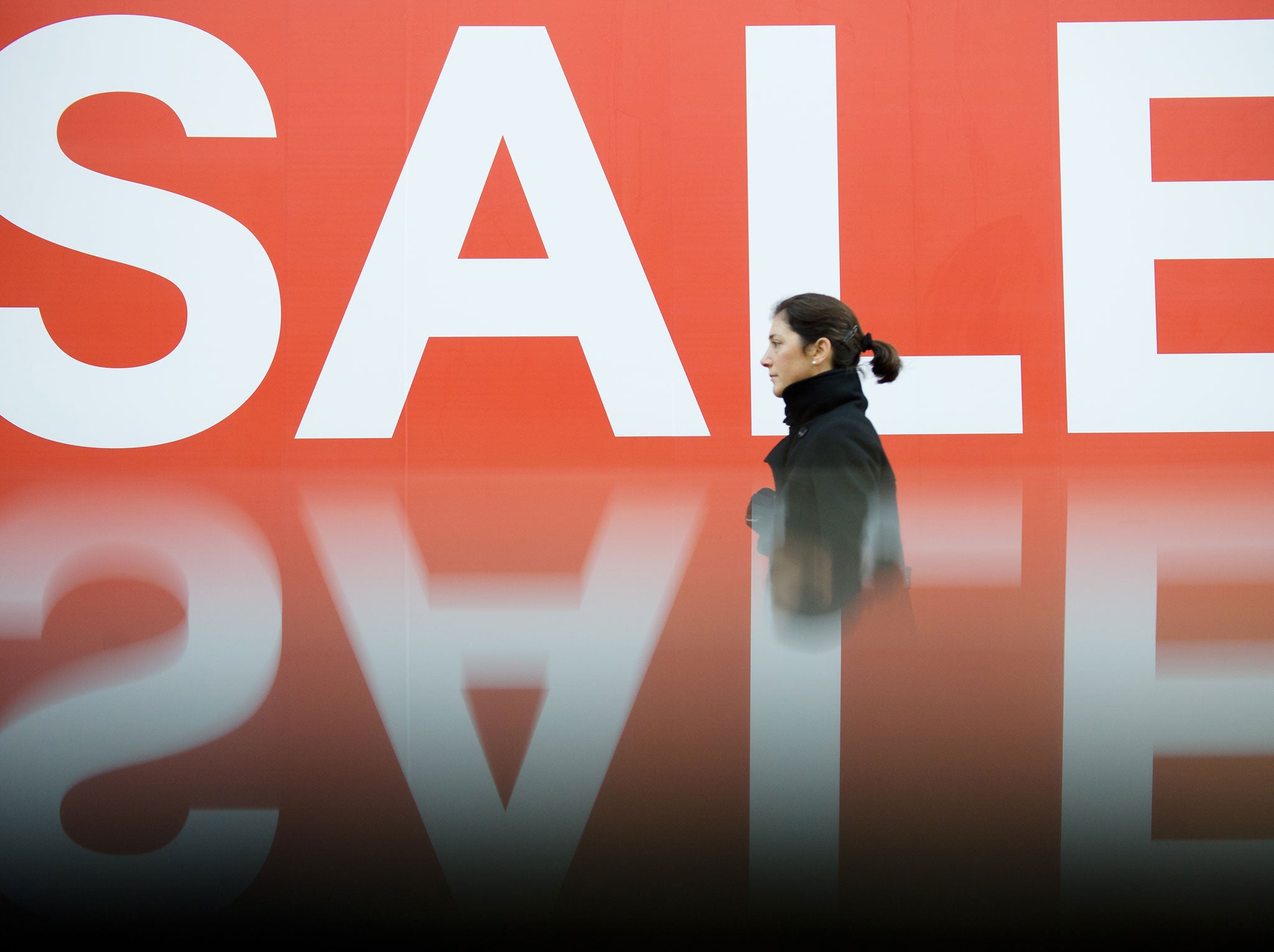 A picture taken using a pre-positioned reflective surface to produce the reflection shows a woman passing a sign advertising an in-store sale in central London on December 16, 2012, less than two weeks before Christmas.