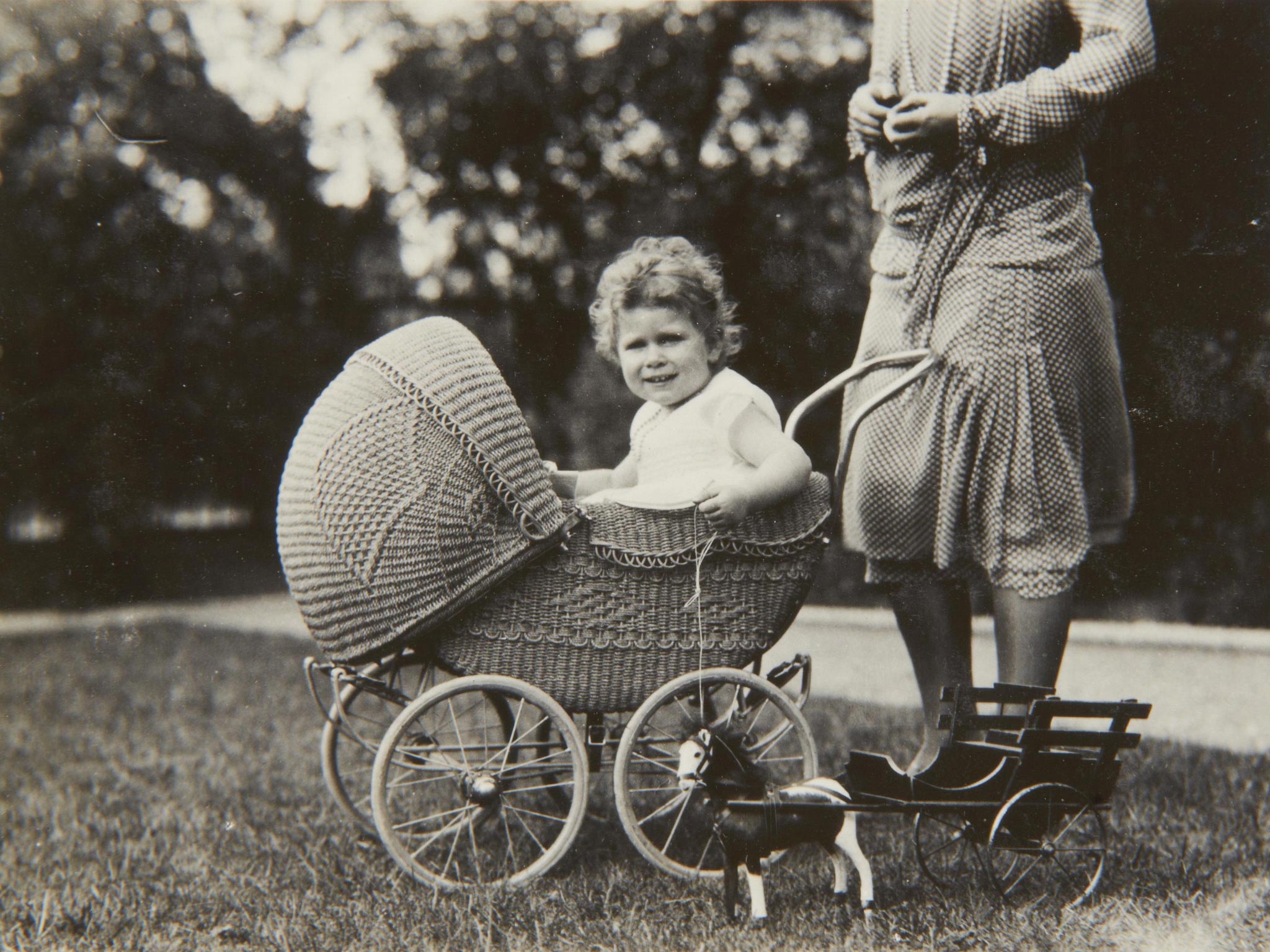 Queen Elizabeth II sitting in a wicker pram with a small toy horse and carriage in 1928