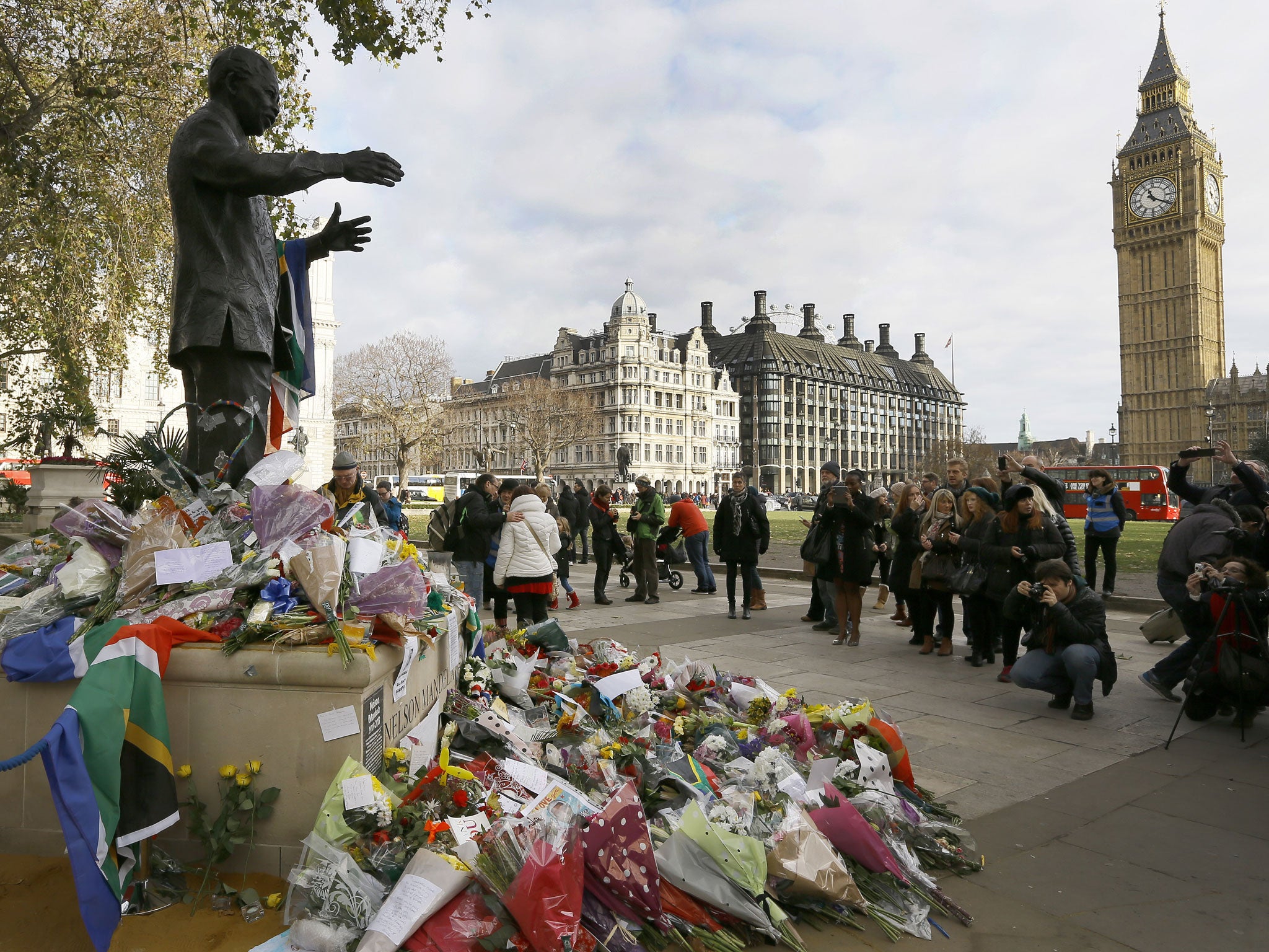 People gather to look at flowers and tributes left at the statue of Nelson Mandela in Parliament Square, London