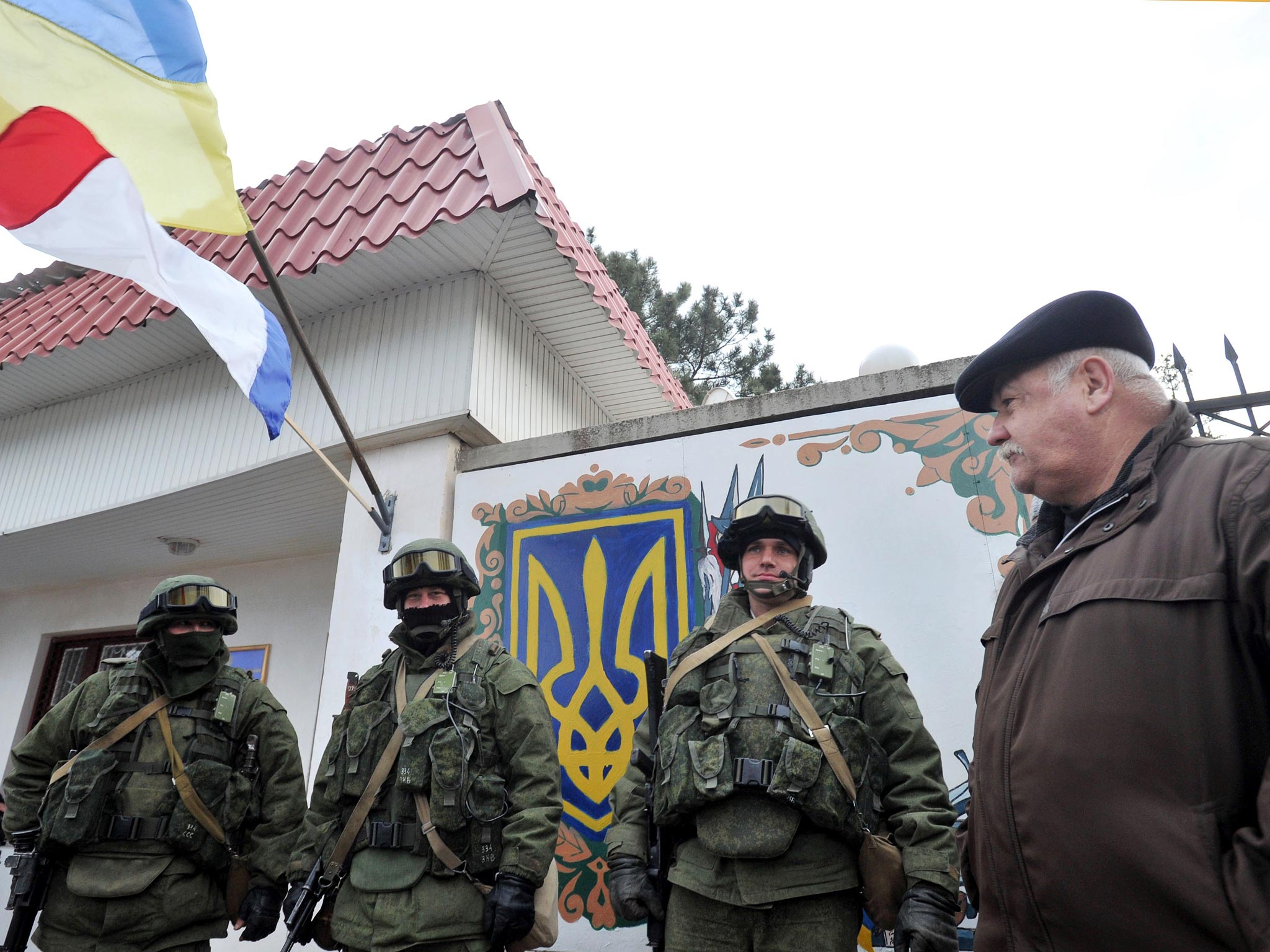 An Autonomous Republic of Crimea and a Ukrainian flag float side by side as a man watches armed men in military fatigues