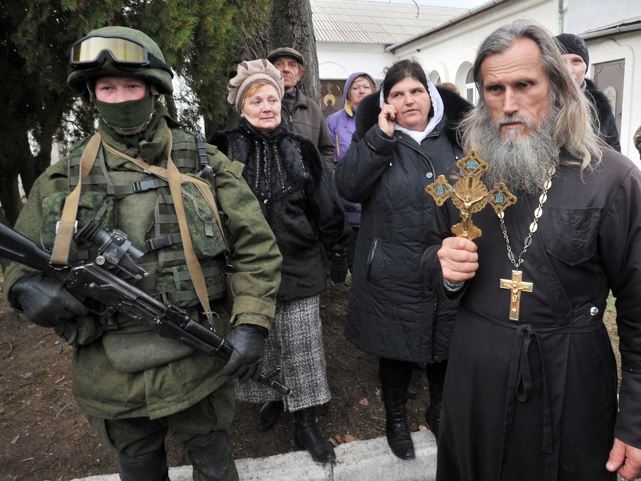 An Orthodox priest holds a crucifix next to one of many armed men in military fatigues blocking access to a Ukrainian border guards base