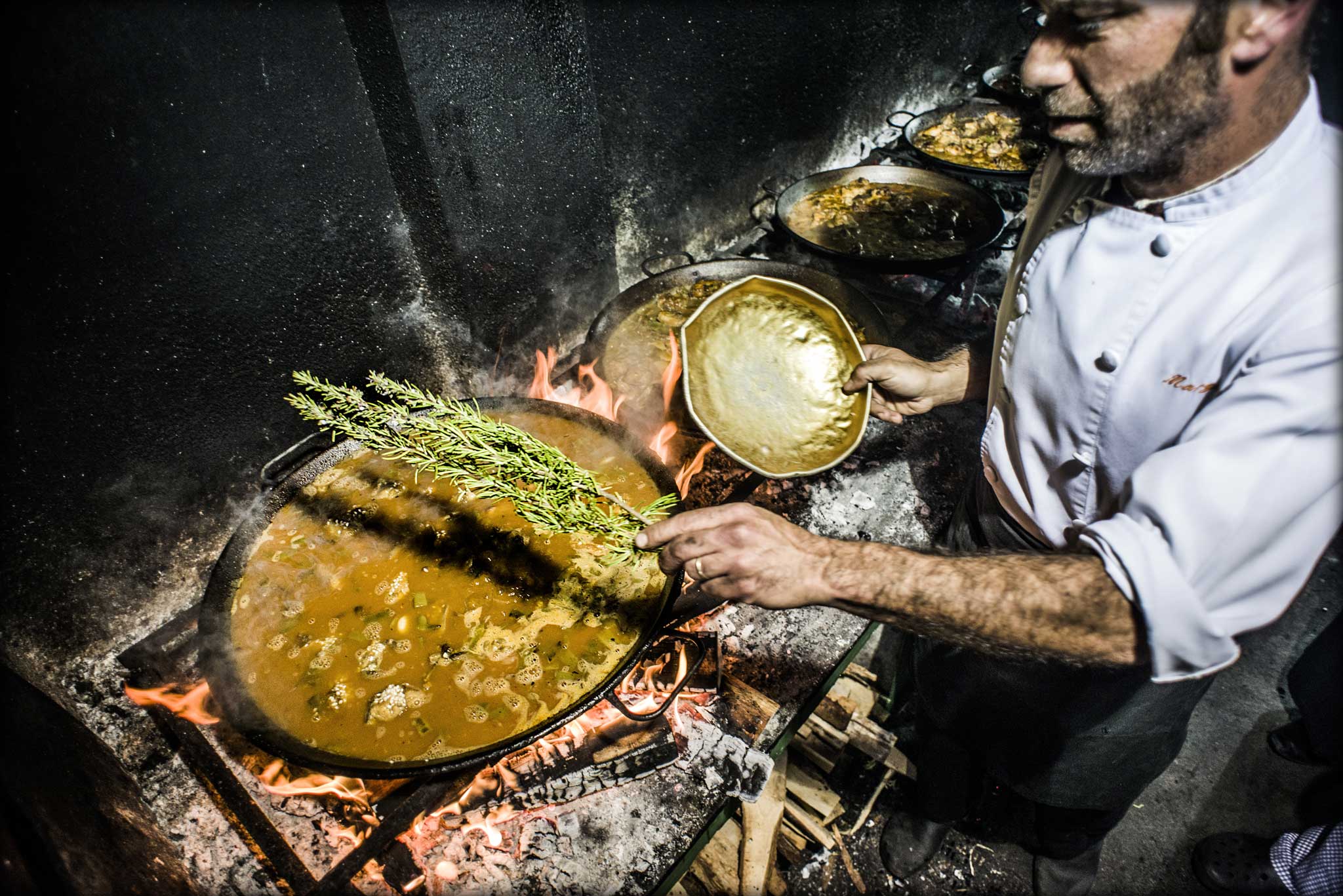 A sweating chef tends each paella, one adding salt, another tossing in a branch of rosemary, a third weighing out rice on an ancient set of scales