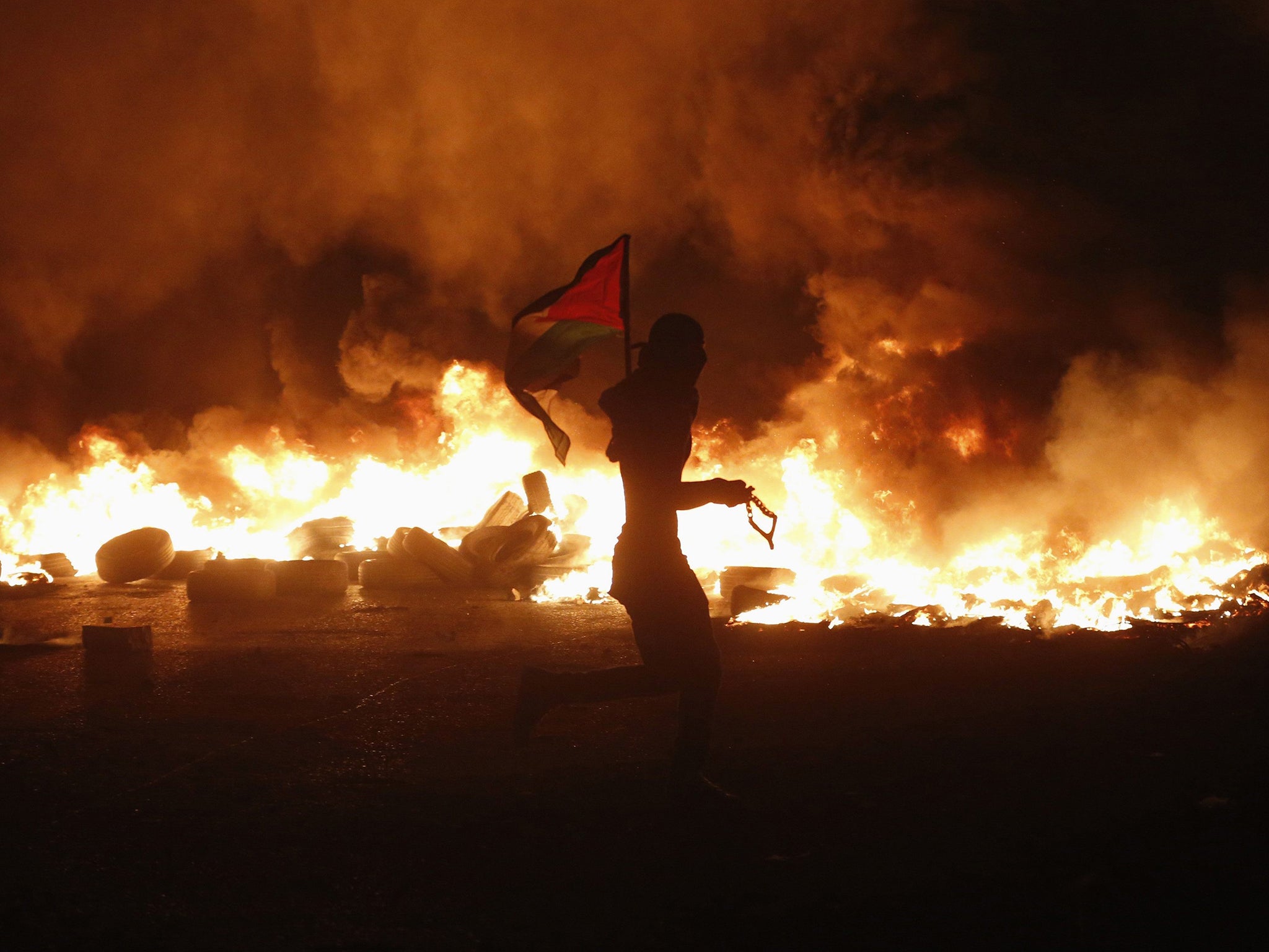 A Palestinian protester holds a Palestinian flag as he runs past burning tyres during clashes with Israeli troops, at a protest against Israeli offensive in Gaza, at Qalandia checkpoint near the West Bank city of Ramallah