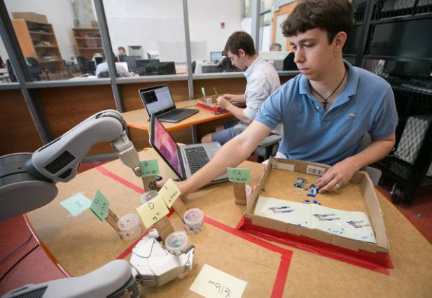 A PR2 robot hands lego pieces to Matthew Gombolay (front) with fellow researcher Giancarlo Sturla in the background. Image credit: Jason Dorfman/CSAIL