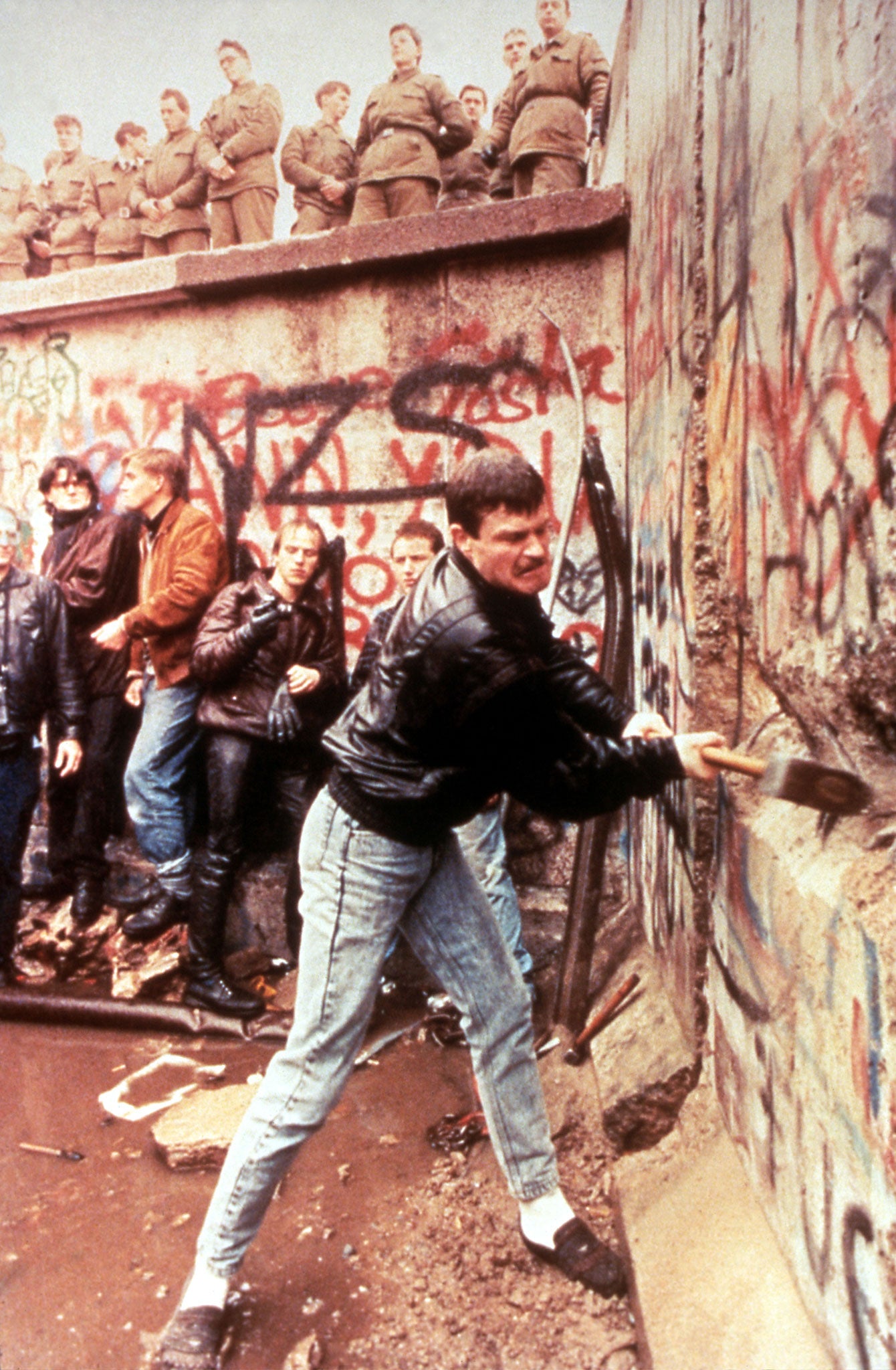 Guards look on impassively as a man sets about the Berlin Wall with a sledgehammer