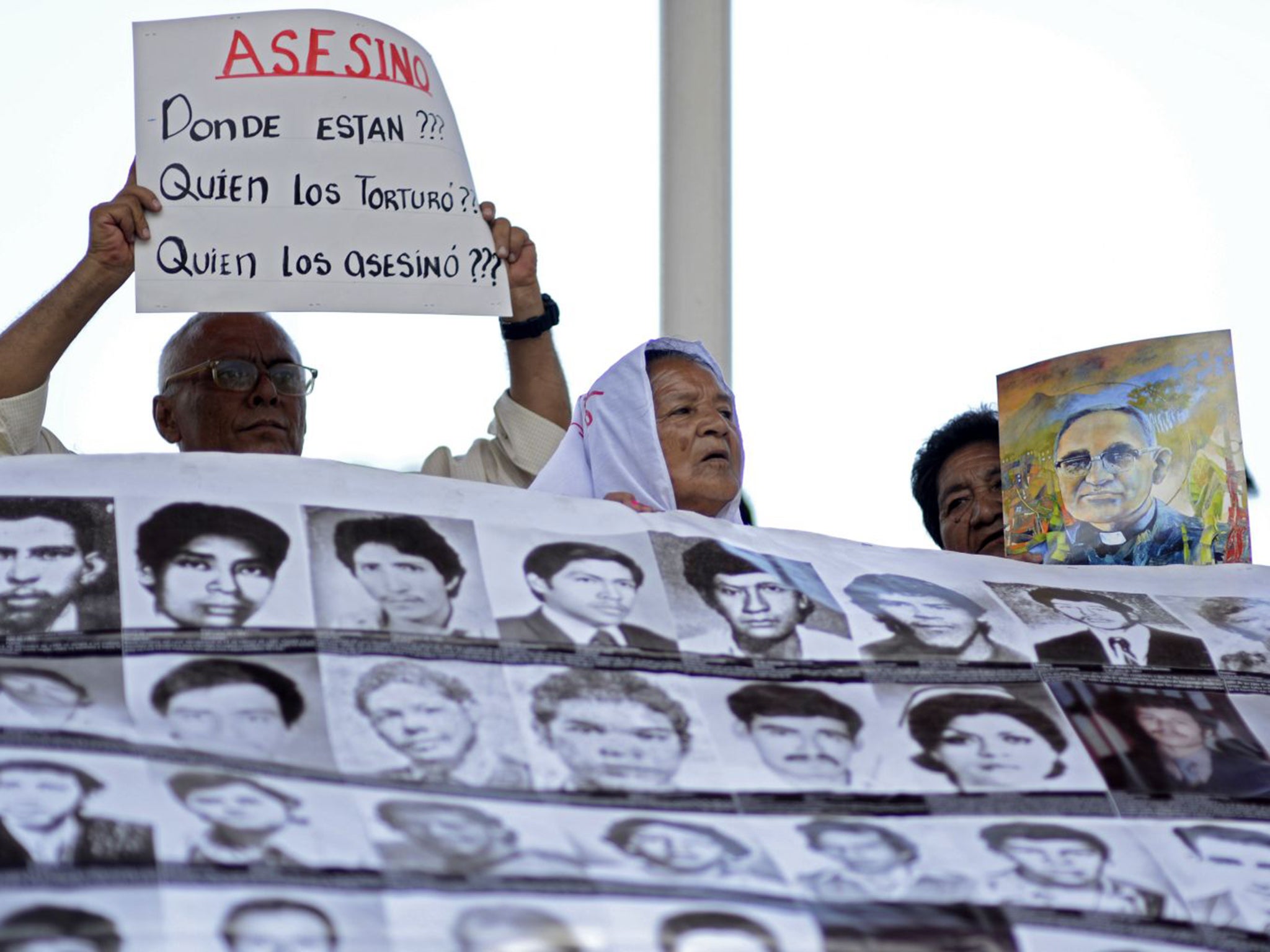 People hold a poster with portraits of disappeared people and a sign reading "Assassin. Who tortured them? Who killed them?"
