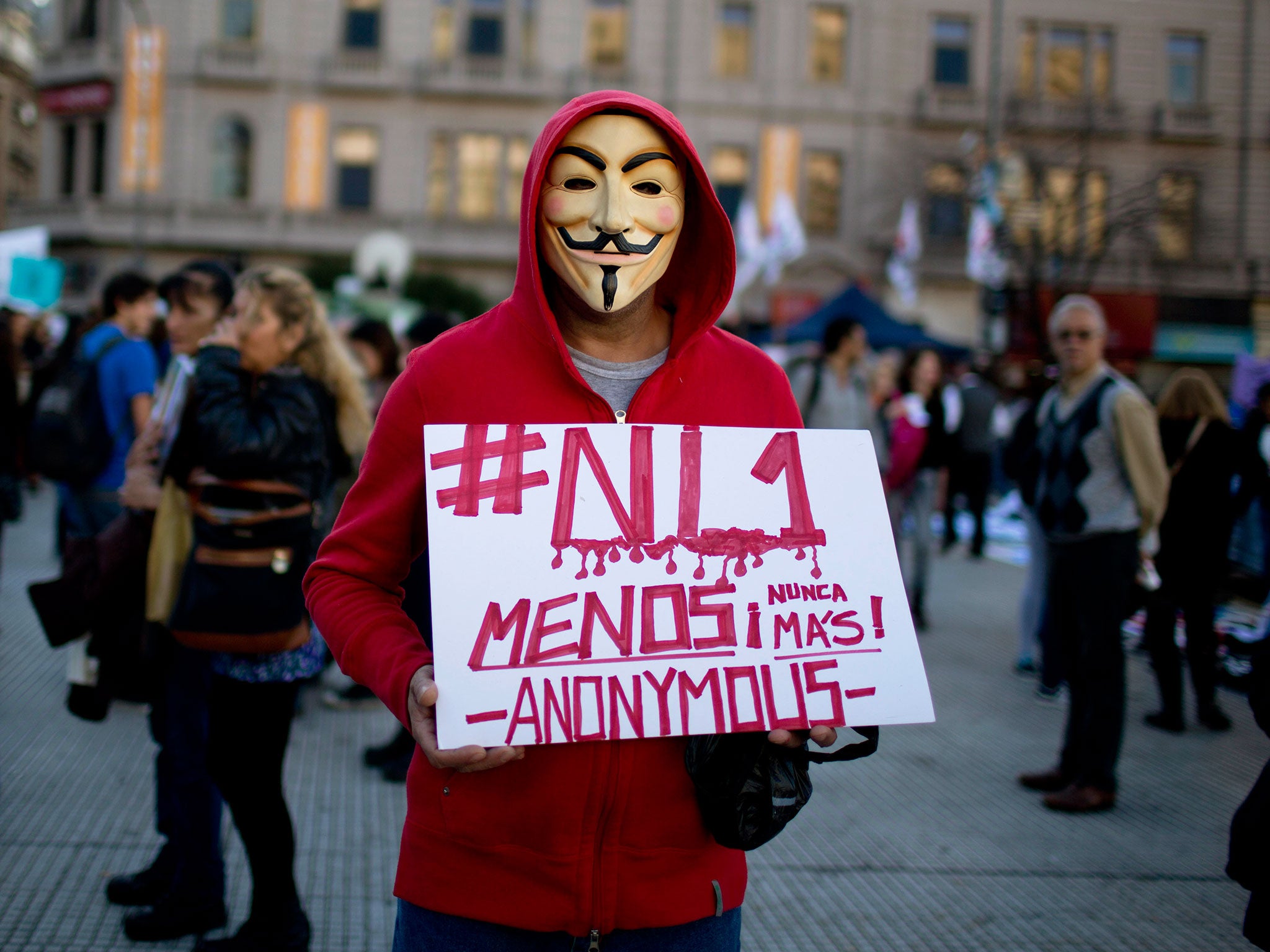 A masked man holds a sign that reads in Spanish "Not one less, never again. Anonymous."