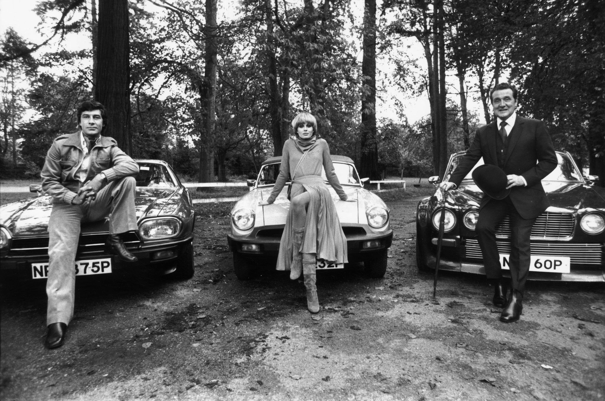 1976: British actors Gareth Hunt, Joanna Lumley and Patrick MacNee, the stars of the television series 'The New Avengers', sitting on the bonnets of their sports cars