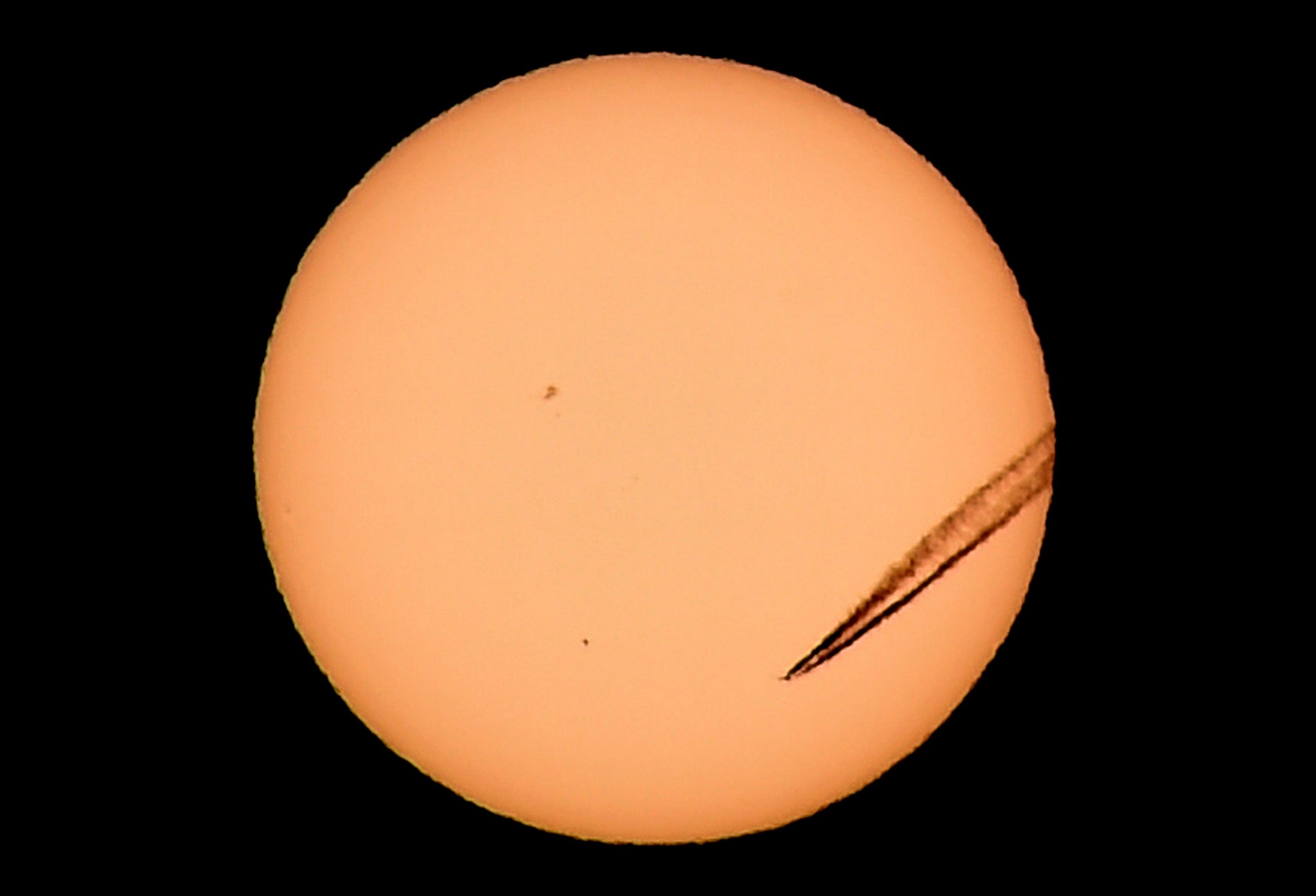 A jet airliner leaves a vapor trail as the planet Mercury is seen in the bottom left transiting across the face of the sun