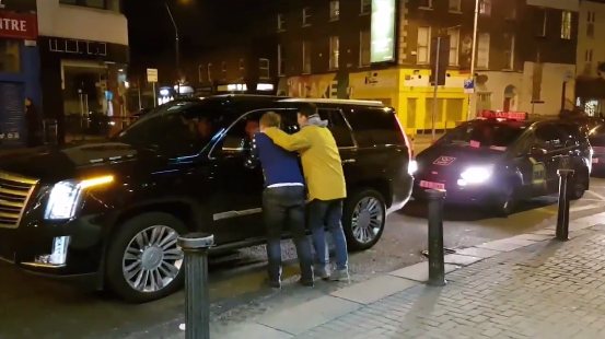 The two men walk over to the car to discover who is in the passenger seat