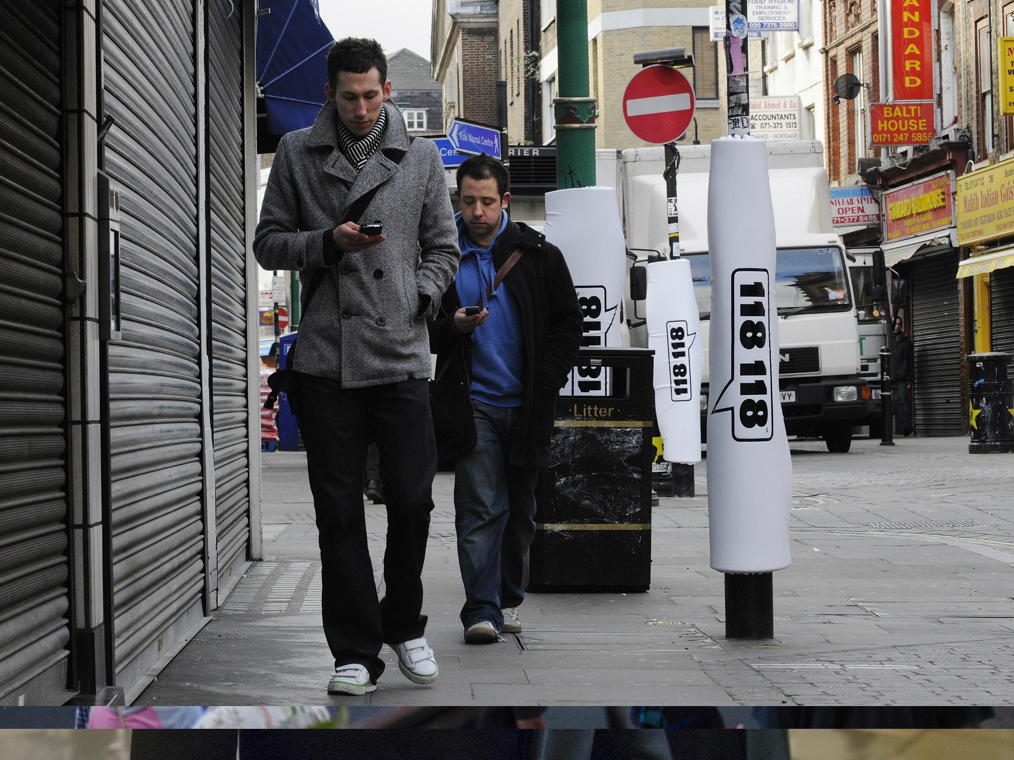 In this photo illustration, two pedestrians walks past padded lamp posts whilst texting in Brick Lane's 'Safe Text' street with padded lamposts.