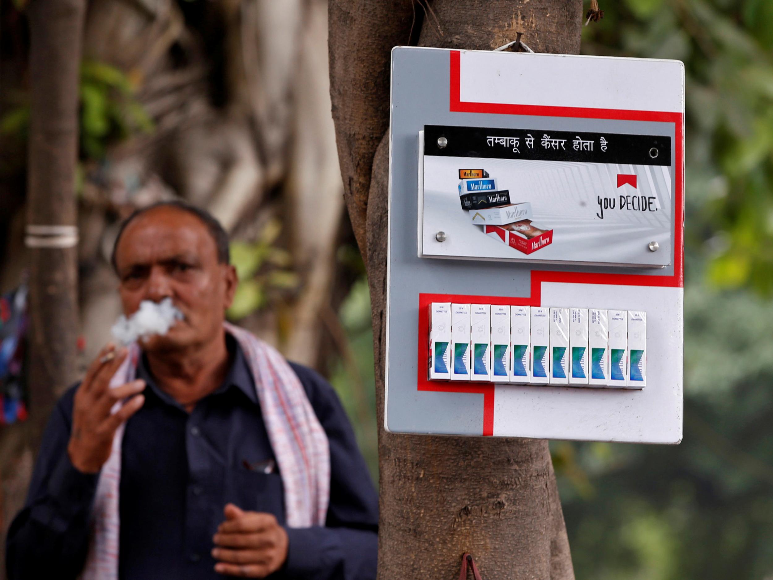 A man smokes next to a cigarette advertisement hung on a tree at a marketplace in New Delhi