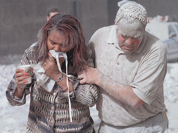 Man covered with ashes assisting a woman walking and holding a particle mask to her face, following the attack