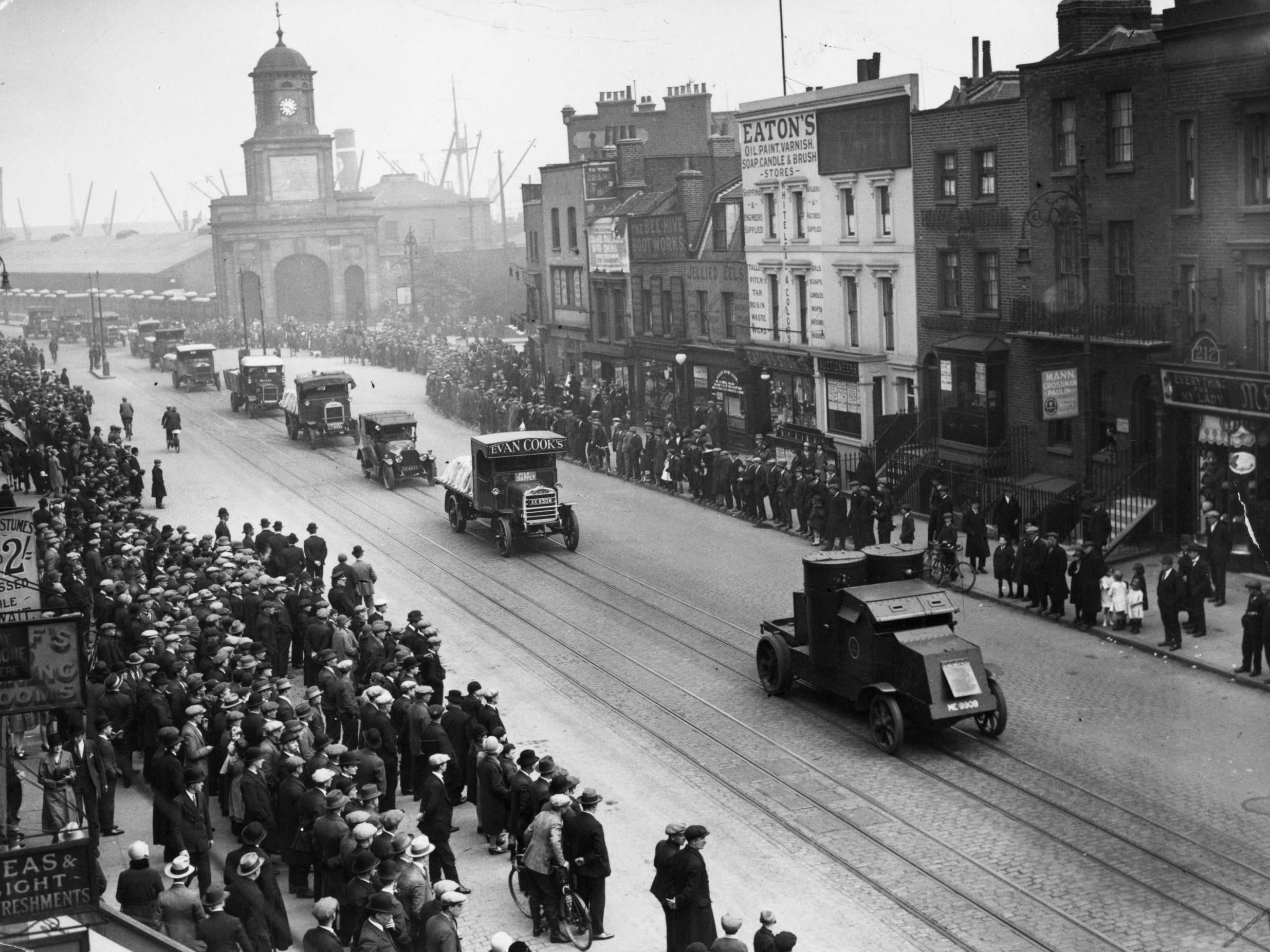 A crowd watches as a food convoy guarded by armoured cars drives down India Dock Road in east London