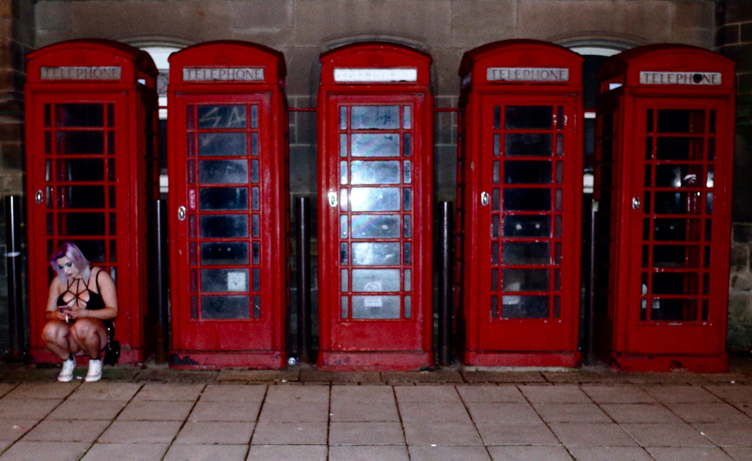 After midnight, New Year’s Eve. A girl looks at her phone and smokes, framed against a line-up of antiquated postcard features of Britain. She’s the most authentic part of the scene, however, a glimpse of modern Britain, while the red phone box belongs to the past