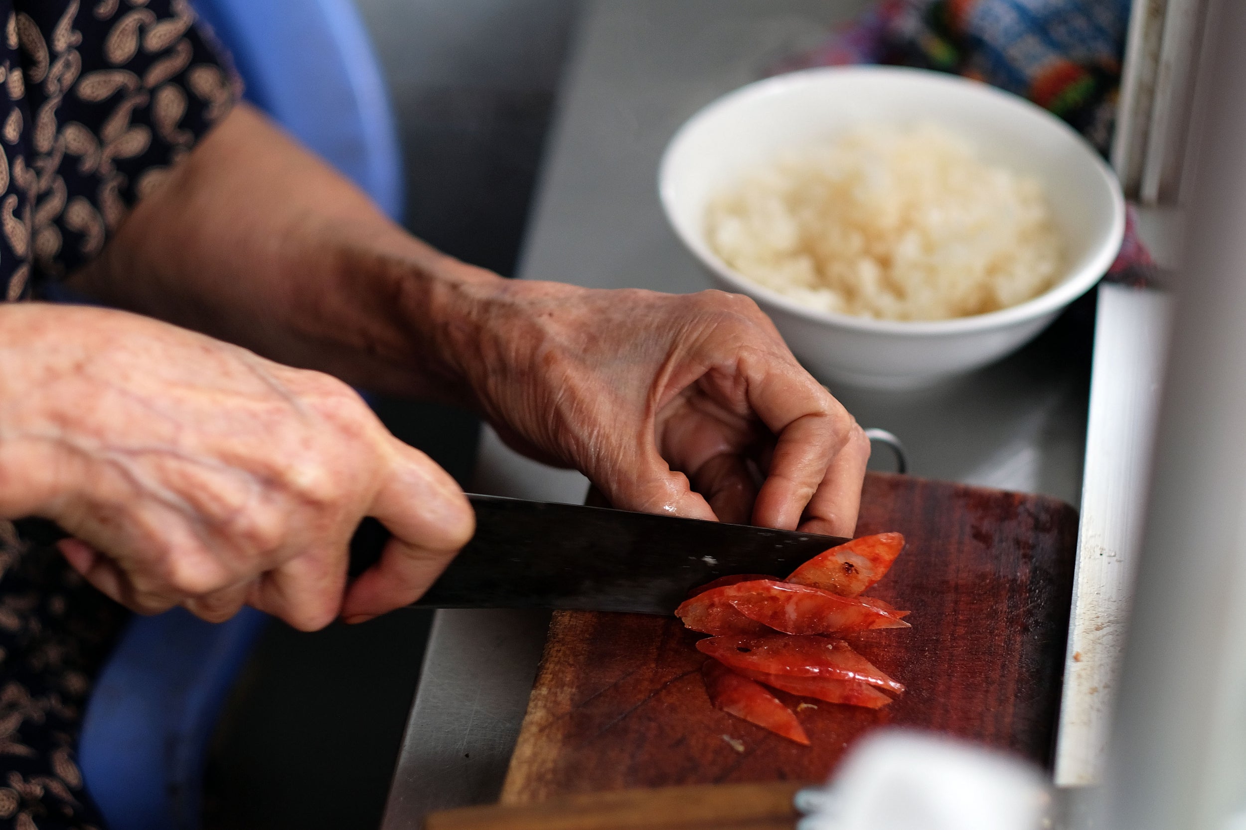 An elderly street cook cuts lap xuong or Chinese sausage, a traditional ingredient when ordering xoi or sticky rice, at a street food stall in Hanoi. Sticky rice is usually served with meat or egg, but there are many variations, such as sticky rice with steamed chicken, sticky rice with mung beans, sticky rice steamed with peanut, and sticky rice with sliced coconut