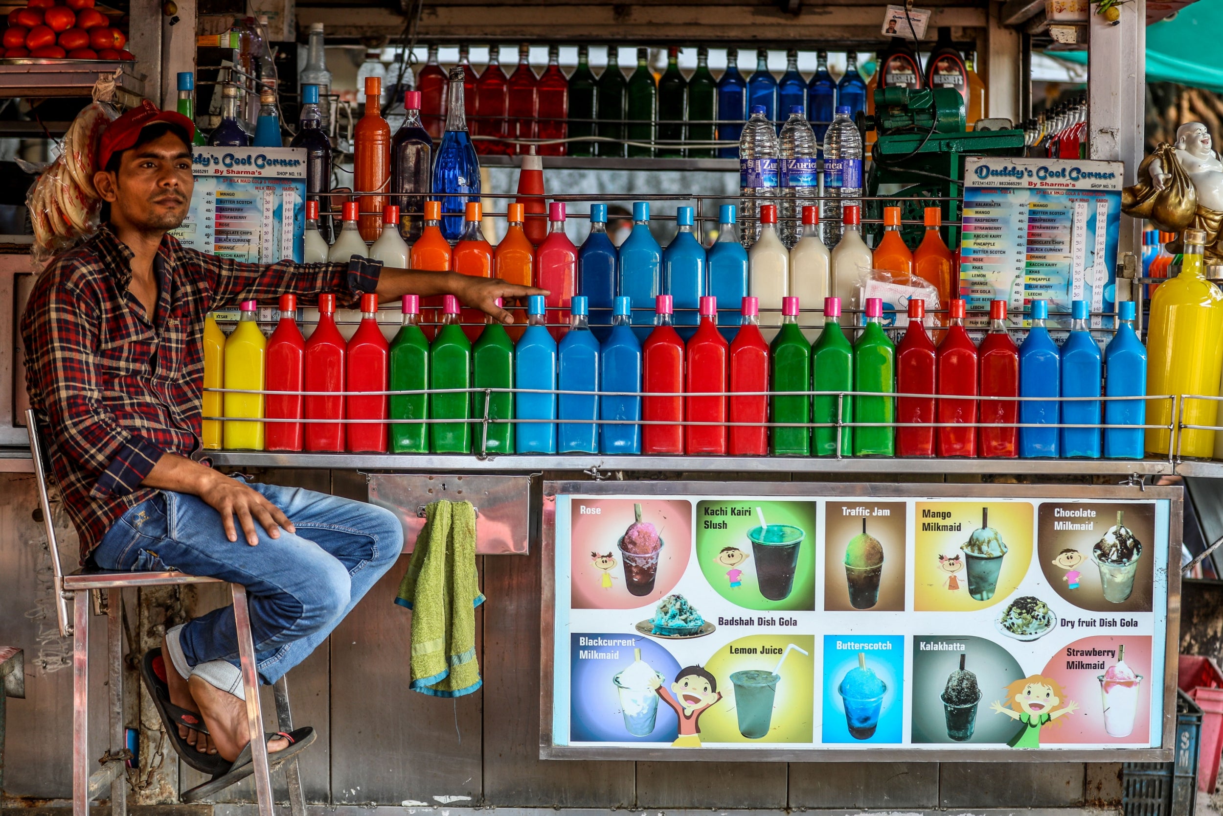 A street food vendor waits for customers seated next to a row of brightly-coloured syrup bottles at his street gola (shaved ice) stall, at Girgaon Chowpaty in Mumbai. Gola or barf ka gola or chuski are the most popular street desserts. The ice-based dessert is made by shaving an ice-block and pouring various flavored syrups on to the snow-like crushed ice