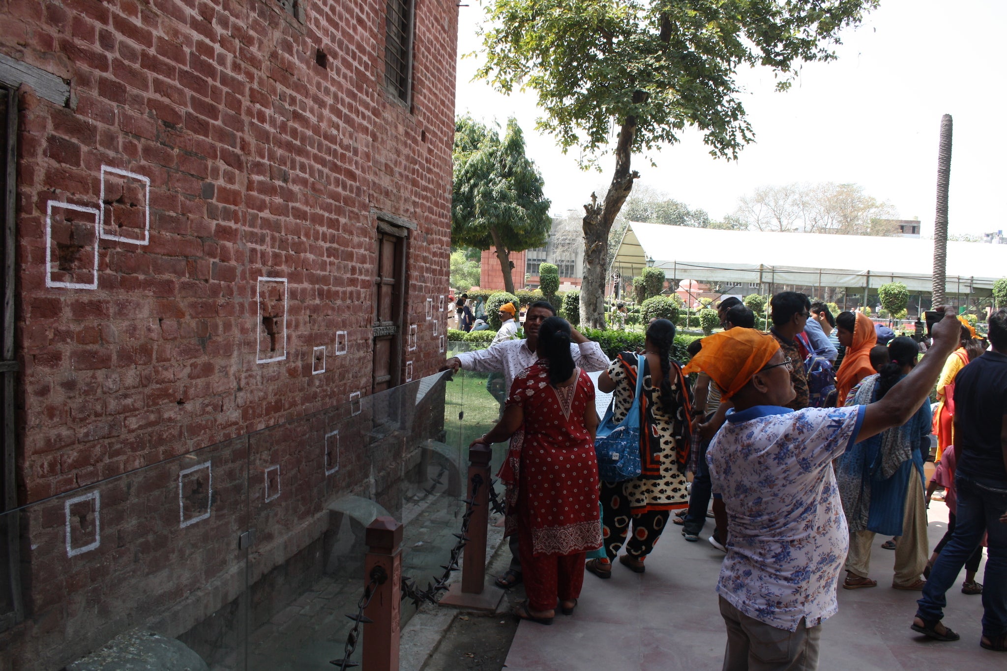 A tourist in a Golden Temple-branded headscarf takes a selfie. Behind him, white paint outline bullet holes partially preserved in the original walls of the garden