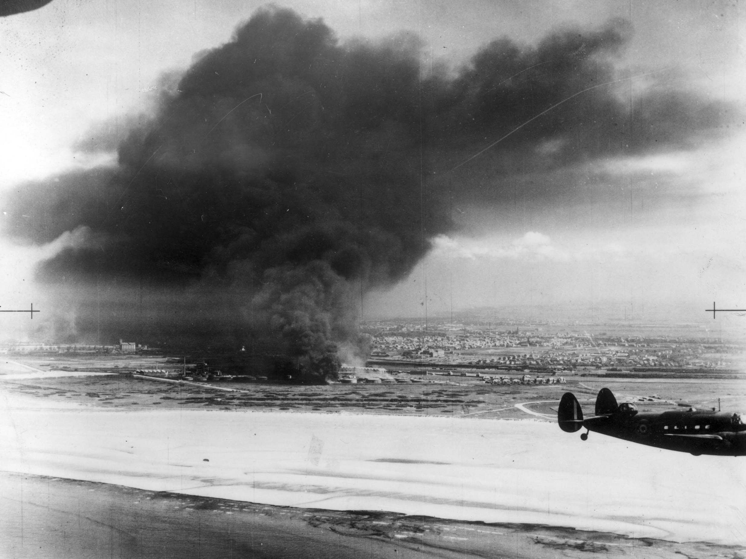 A Coastal Command Lockheed Hudson flying past burning oil tanks on Dunkirk beach