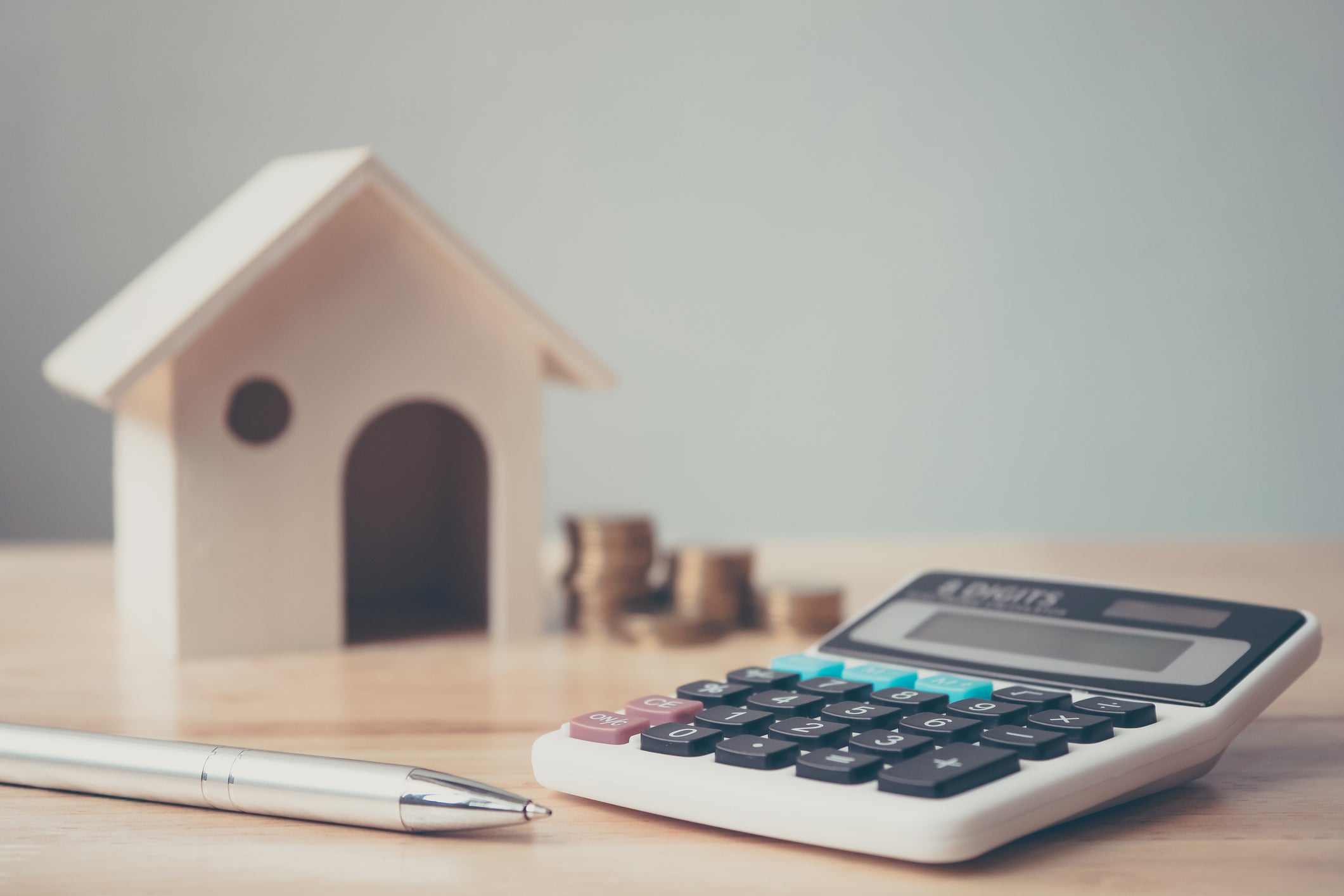 Calculator with wooden house and coins stack and pen on wood table.