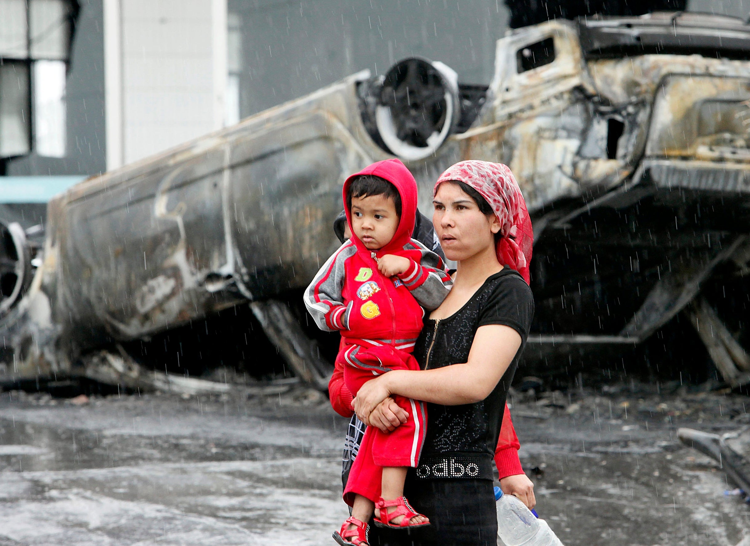 An Uighur woman and child walk past a burned car following riots in Urumqi 10 years ago