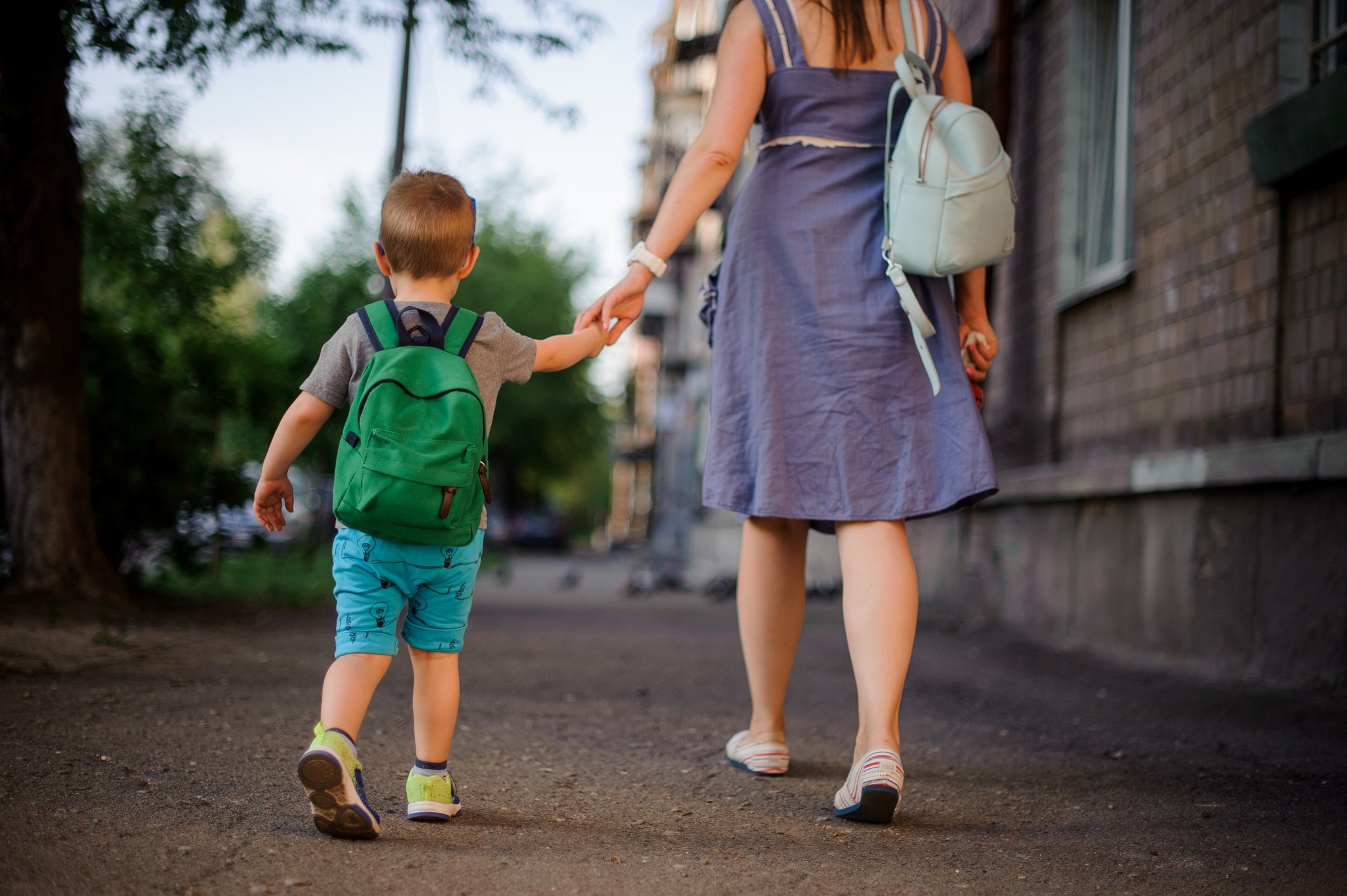 Back view of mother walking down the street with a little son with a backpack on summer day