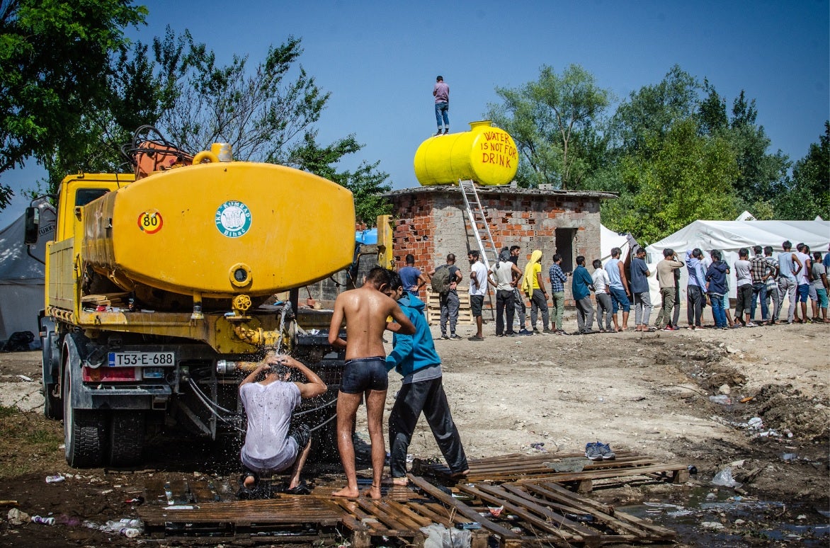 Residents of the camp wait in line for food distributed from Bihać Red Cross, while men wash themselves at the back of a water truck.