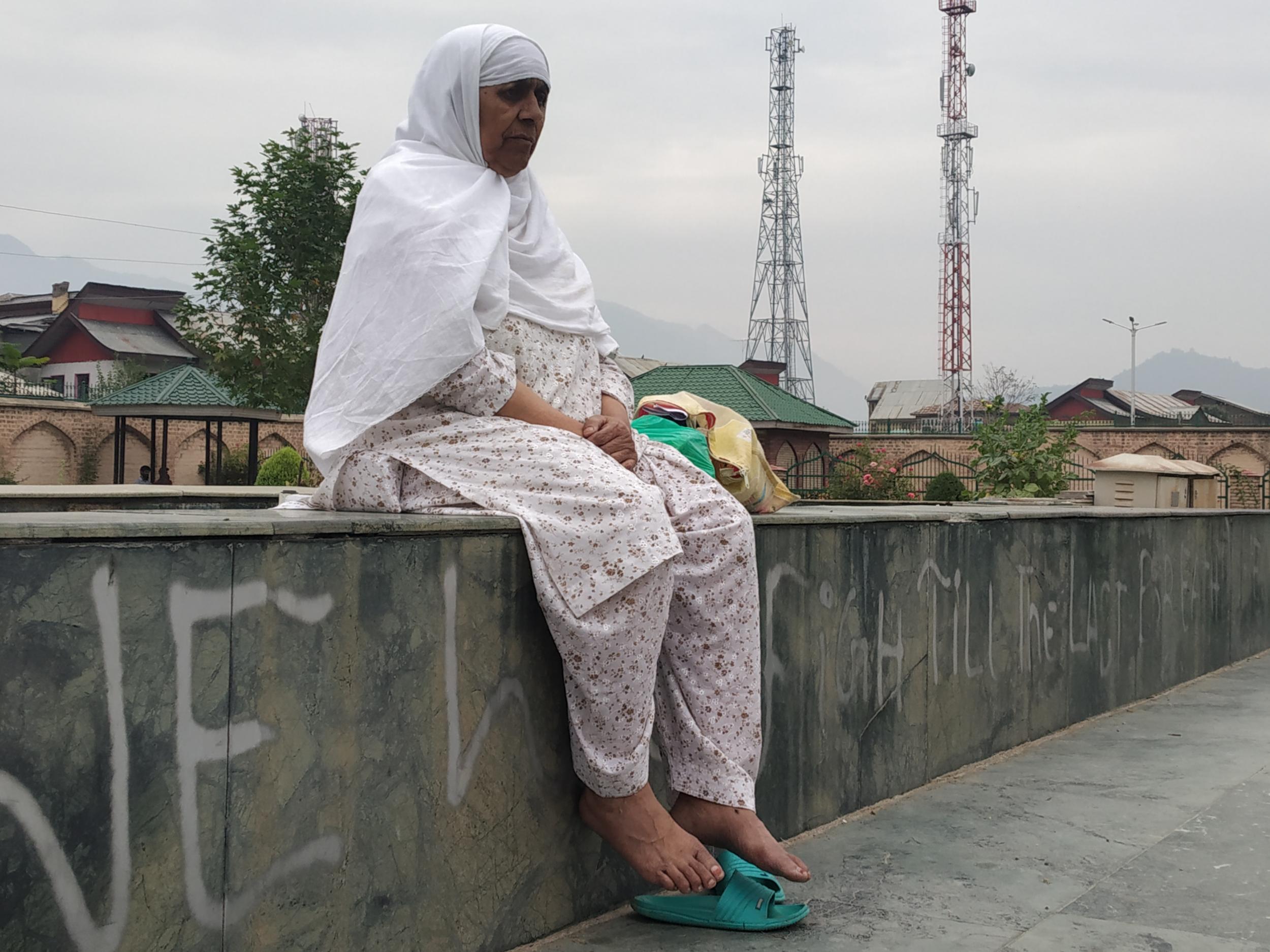 Mugli sits on a wall waiting to see her son. The wall's graffiti reads: 'We Will Fight Till The Last Breath. We Want Freedom'