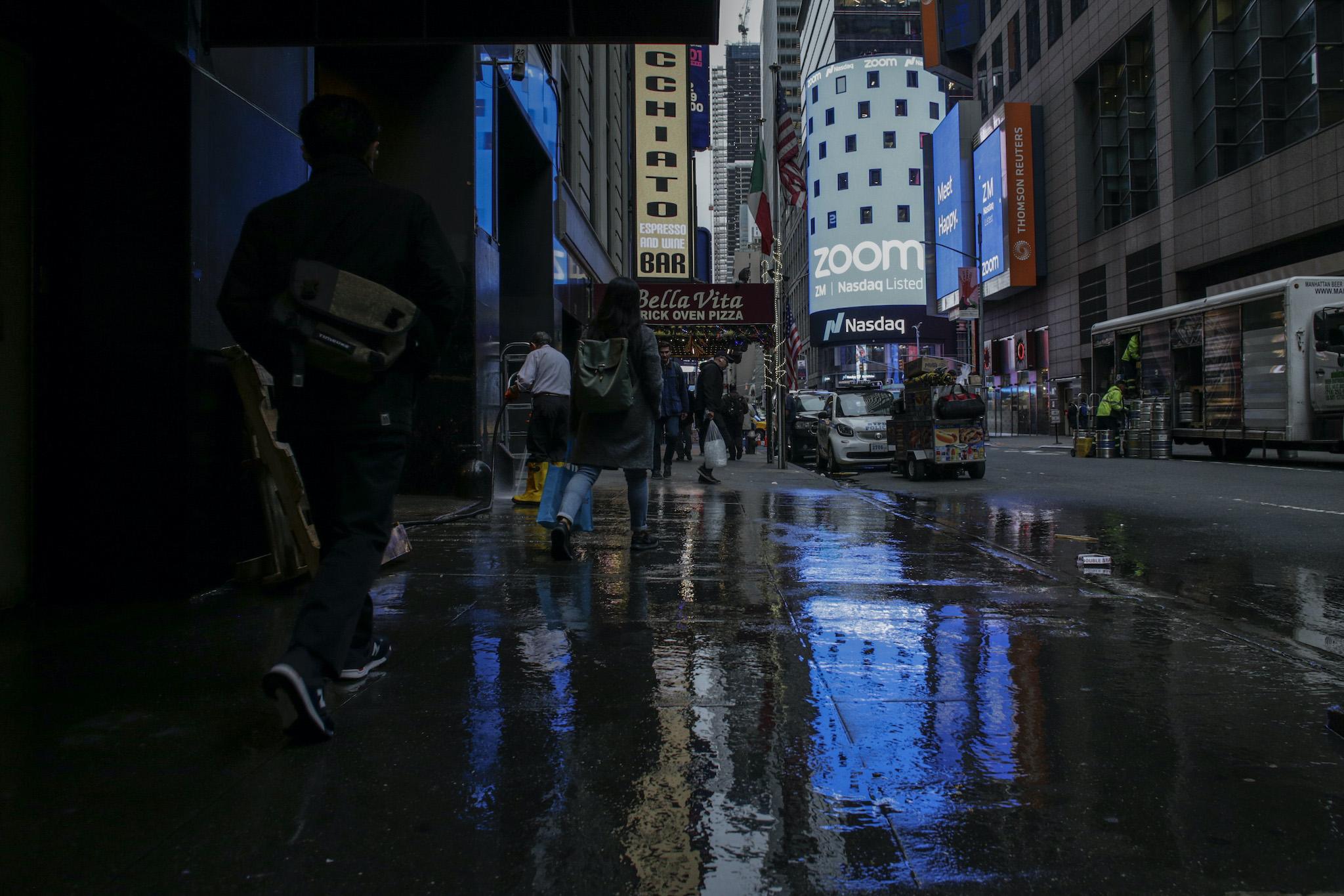 People pass walk by the Nasdaq building as the screen shows the logo of the video-conferencing software company Zoom after the opening bell ceremony on April 18, 2019 in New York City