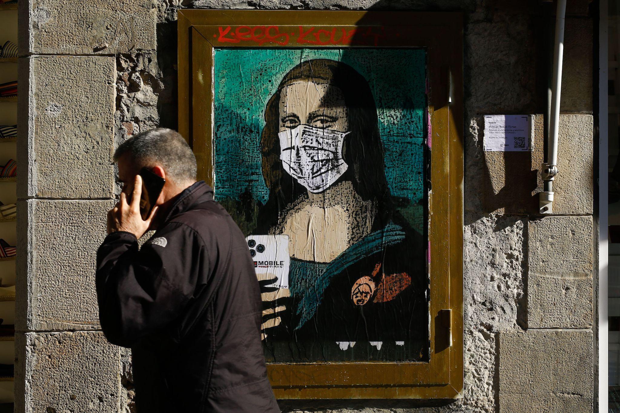 A man holding his mobile phone walks past a poster by Italian urban artist Salvatore Benintende aka "TVBOY" depecting Leonardo da Vinci's Mona Lisa wearing a protective facemask and holding a mobile phone reading "Mobile World Virus" in a street of Barcelona