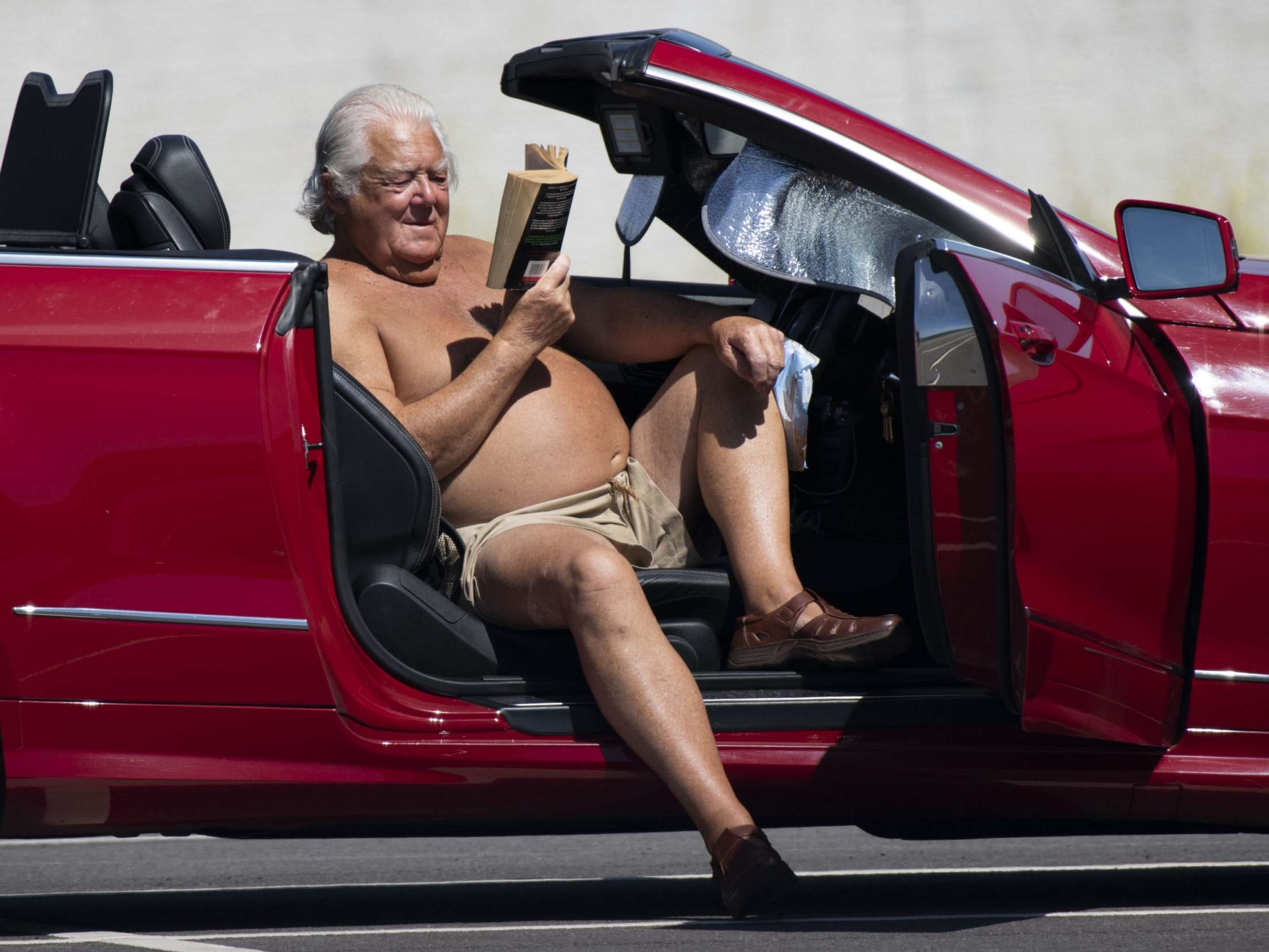 A man reads a book while sitting in the driver seat of his car in the sun in the car park of a Trago Mills department store in Merthyr Tydfil, United Kingdom