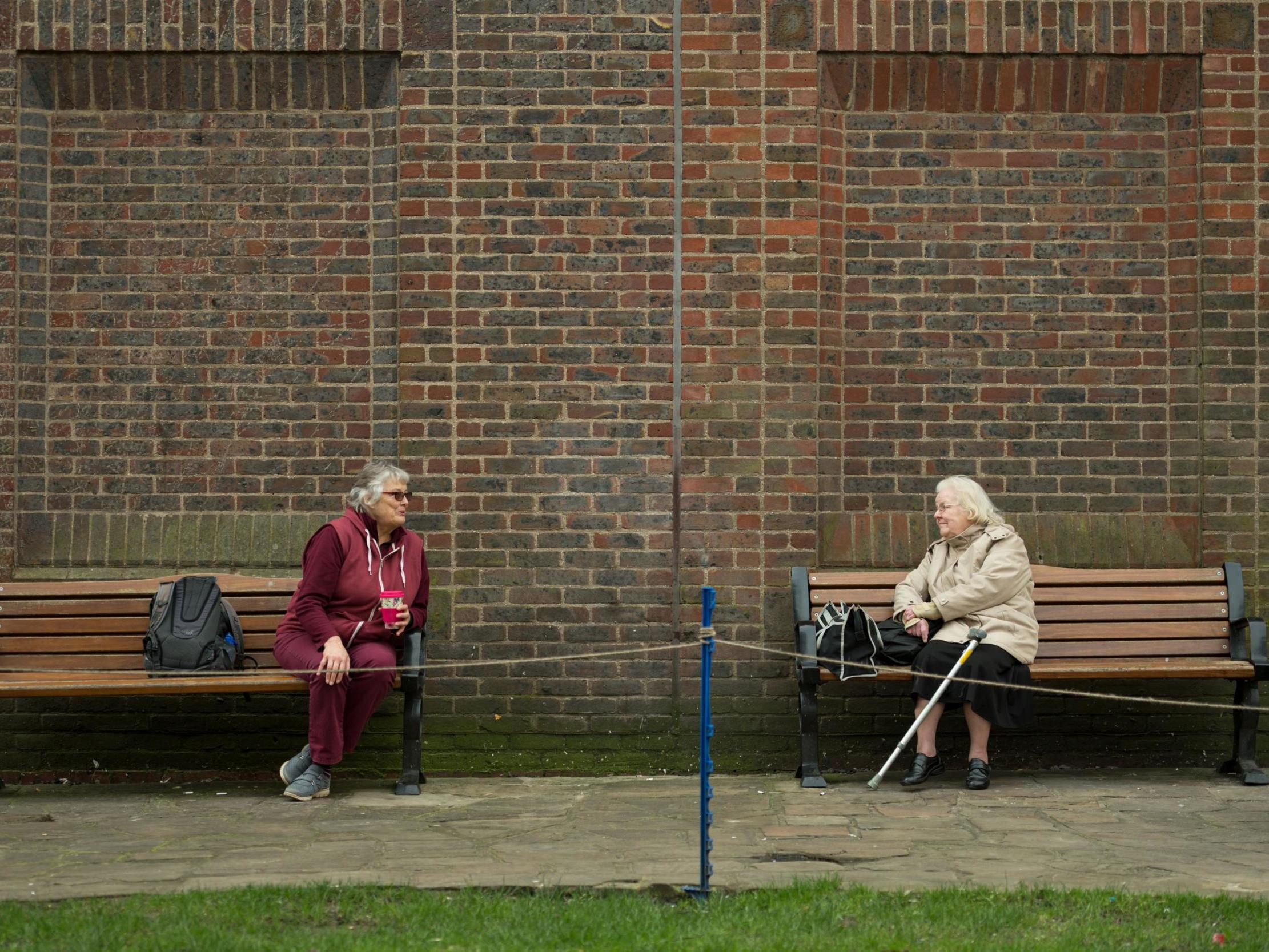 Two women observe social distancing measures as they speak to each other from adjacent park benches