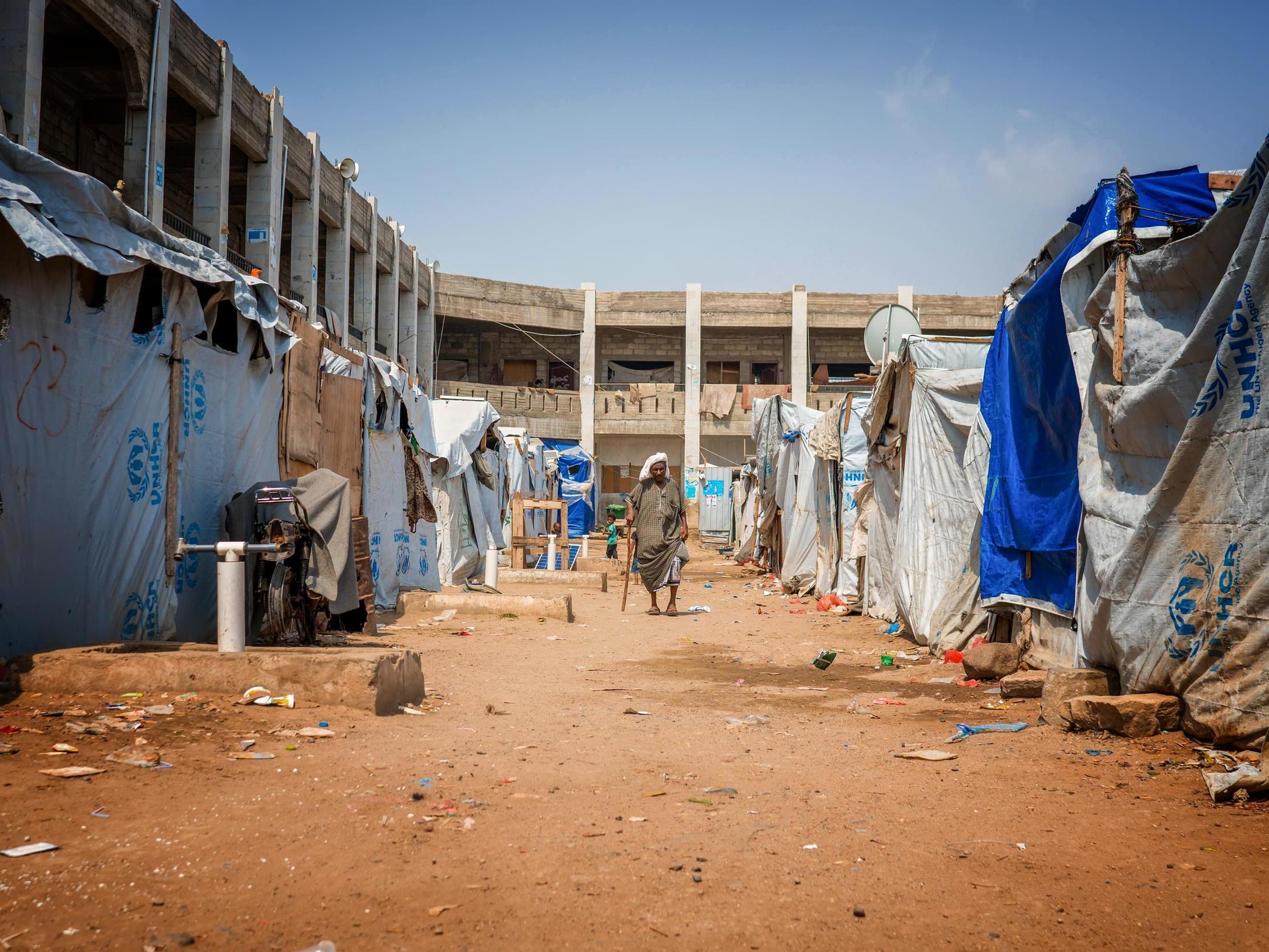 A man with a walking stick in the yard between newly built tents and an unfinished school building that is being used as a shelter